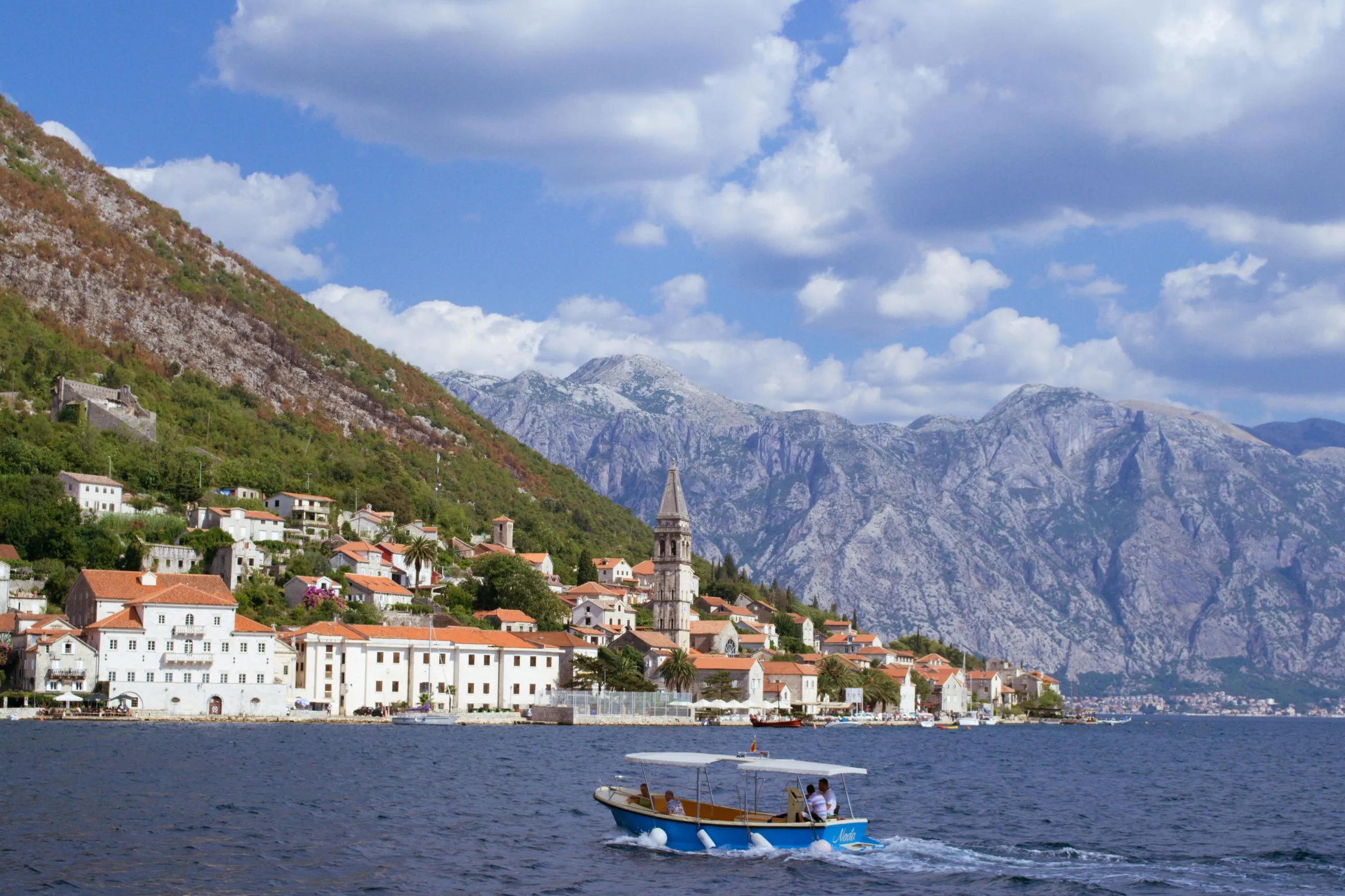 A small boat sailing in the Bay of Kotor, Montenegro, with the picturesque town of Perast and dramatic mountains in the background.