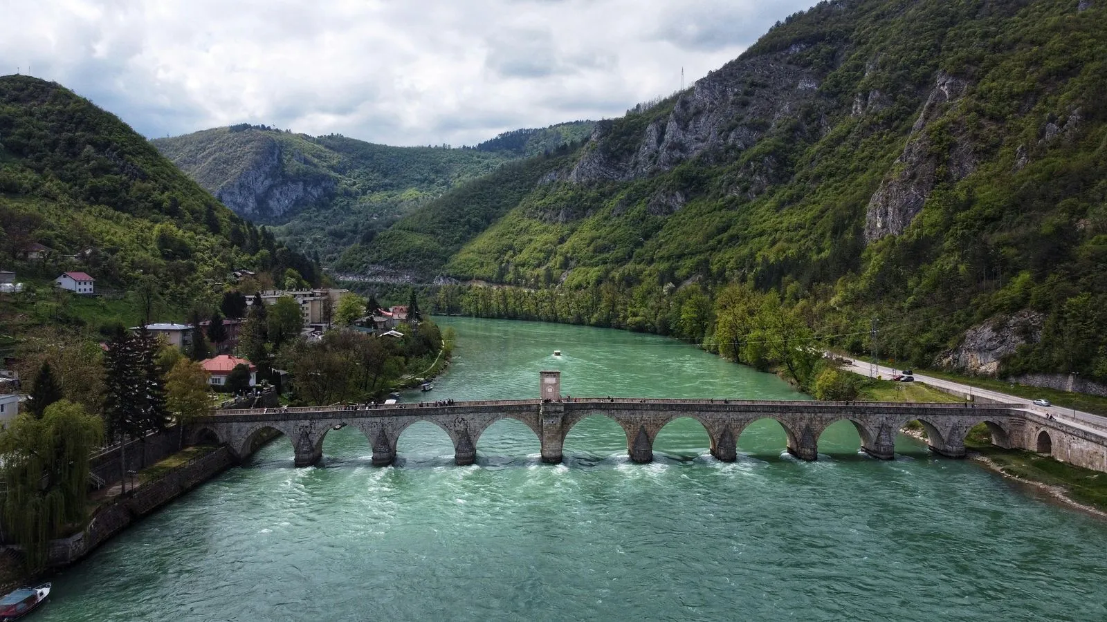 Aerial view of the Mehmed Paša Sokolović Bridge in Višegrad, Bosnia, surrounded by lush green hills and turquoise water.