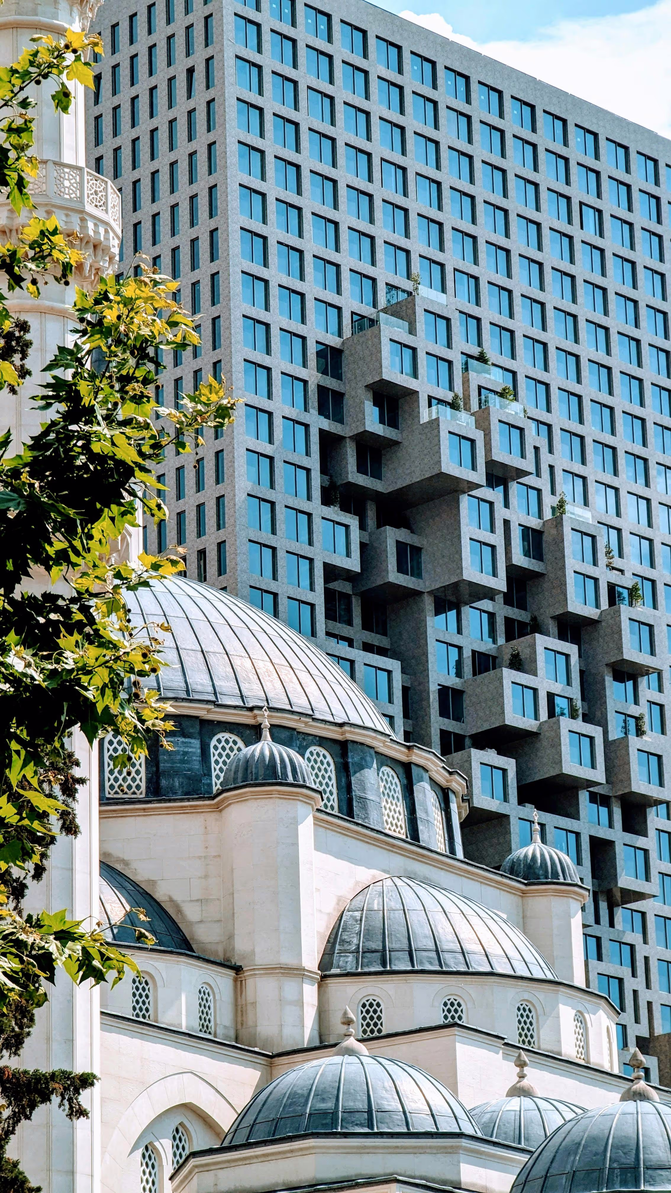 A modern geometric high-rise building behind an Ottoman-style mosque with domed rooftops in Tirana, Albania.
