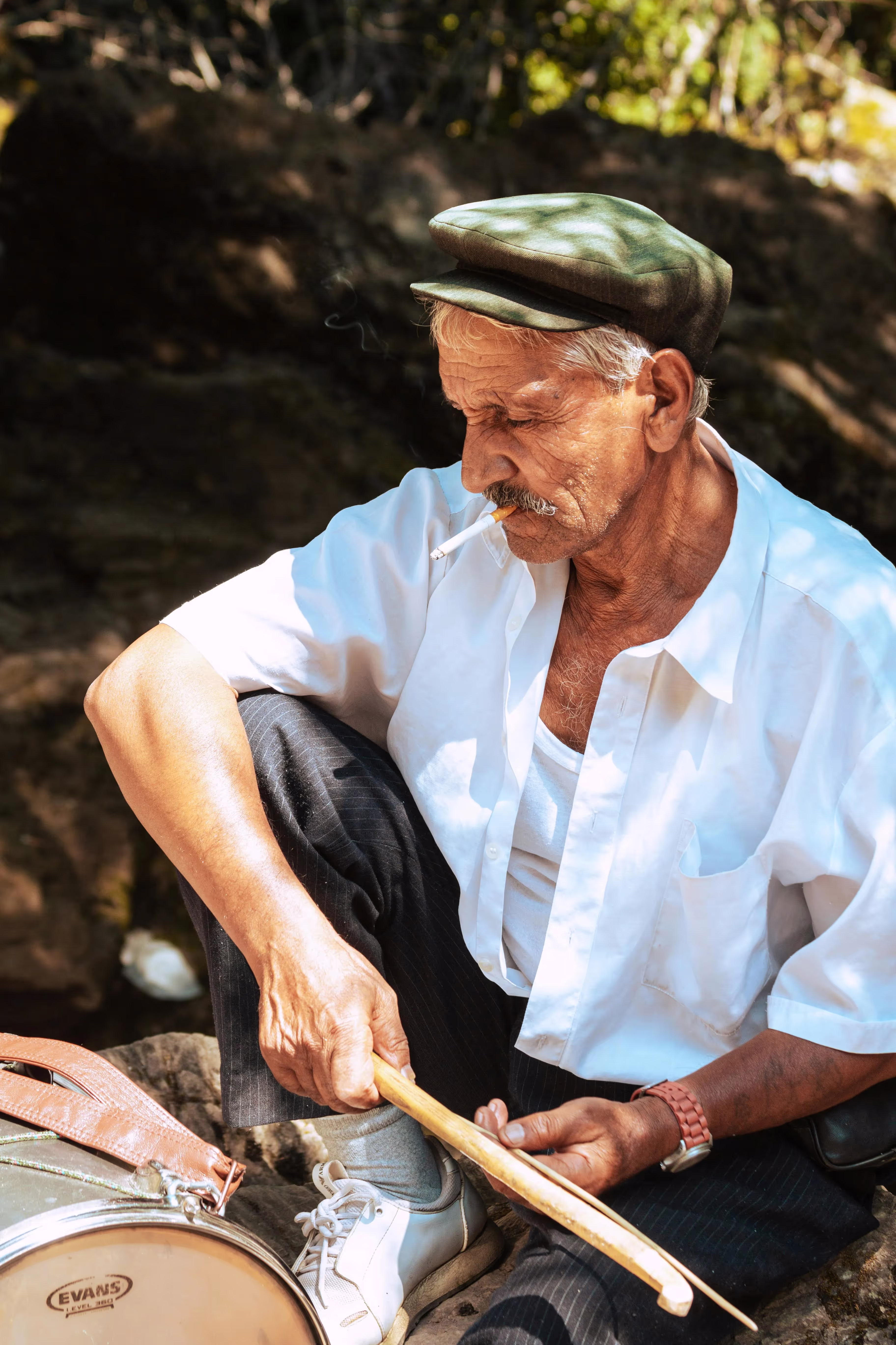 Elderly man in a white shirt and cap playing drums outdoors in Albania, smoking a cigarette.