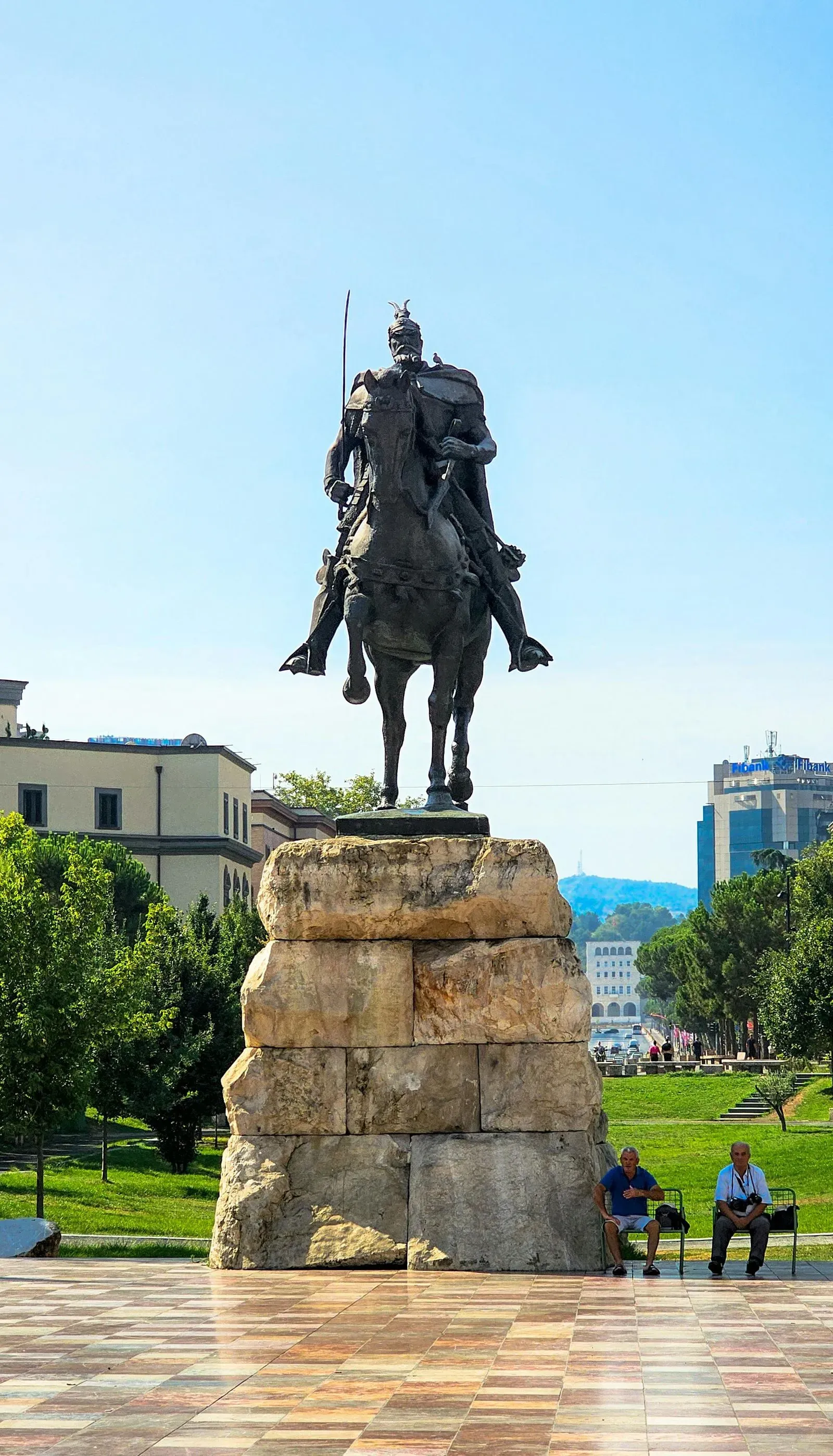 The equestrian statue of Skanderbeg in Tirana, Albania, standing on a stone pedestal with two elderly men seated nearby.