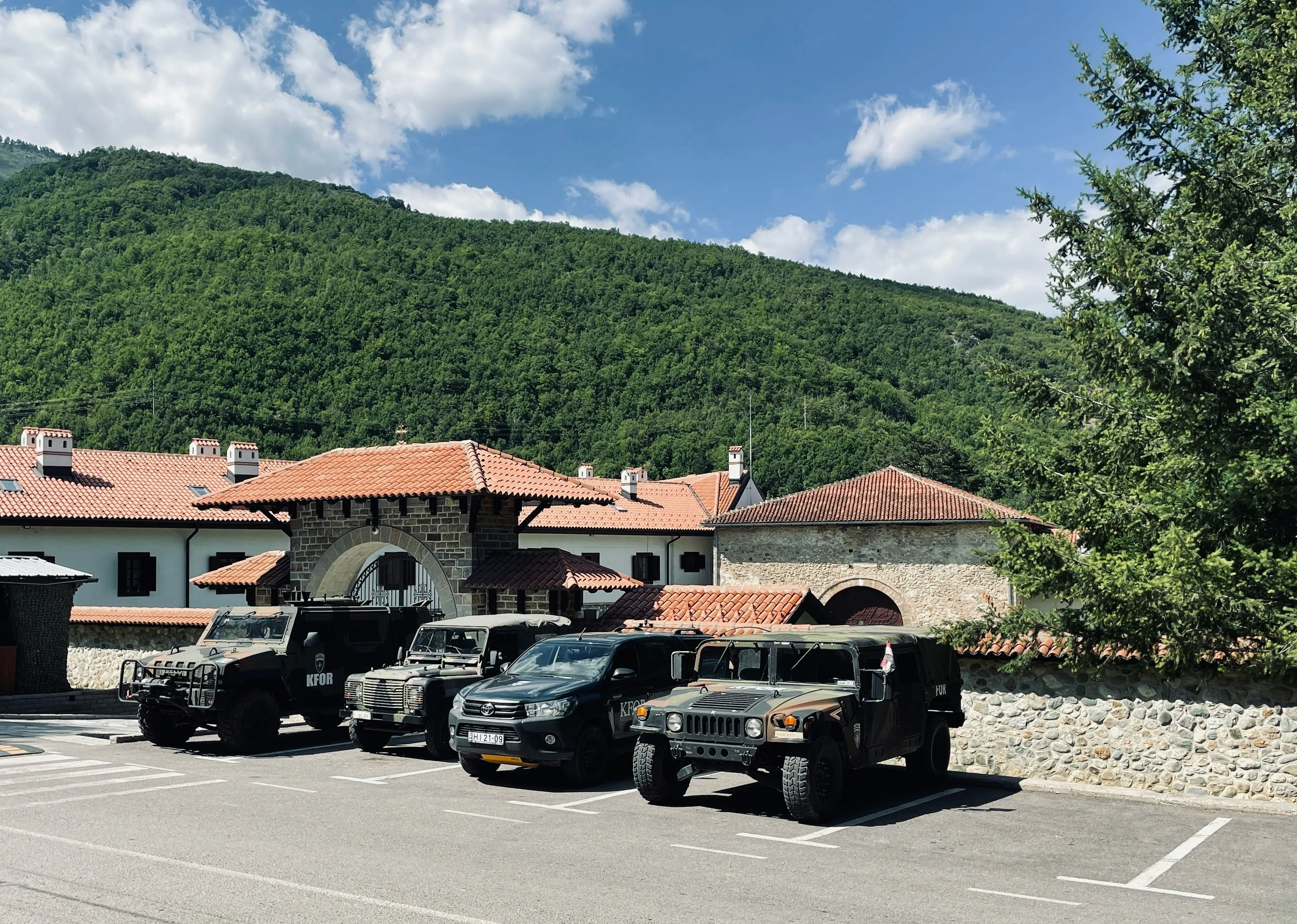 Military vehicles parked outside Visoki Dečani Monastery in Kosovo, surrounded by lush green mountains.