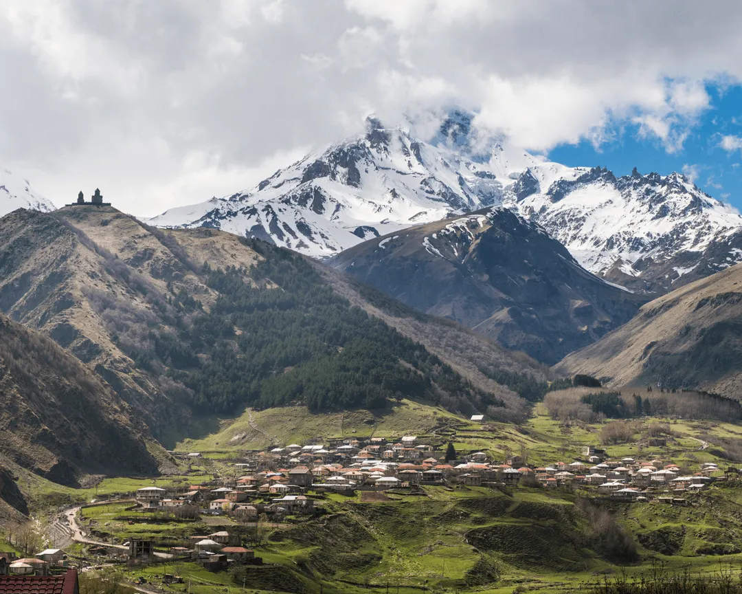Snow-capped Mount Kazbegi with the Gergeti Trinity Church perched on a hill, overlooking a small village.