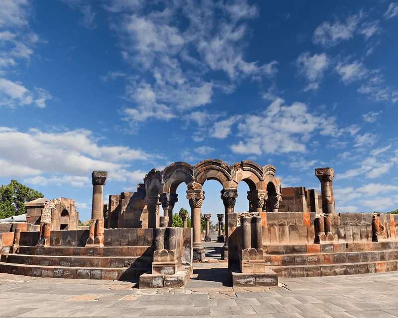 The ruins of Zvartnots Cathedral in Armenia, featuring ornate stone arches under a bright blue sky.