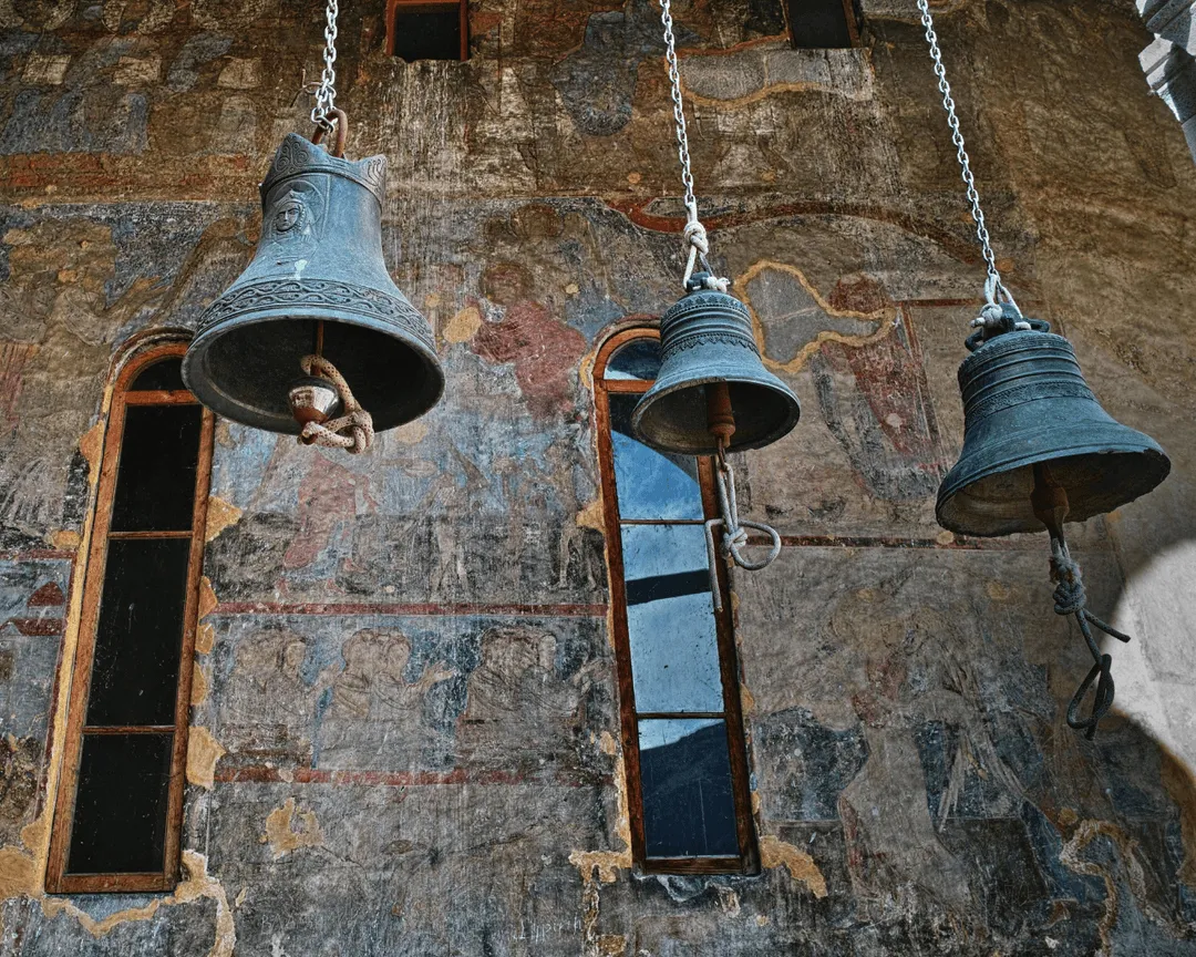 Three antique church bells hanging in front of a weathered fresco at a historic monastery in Georgia.