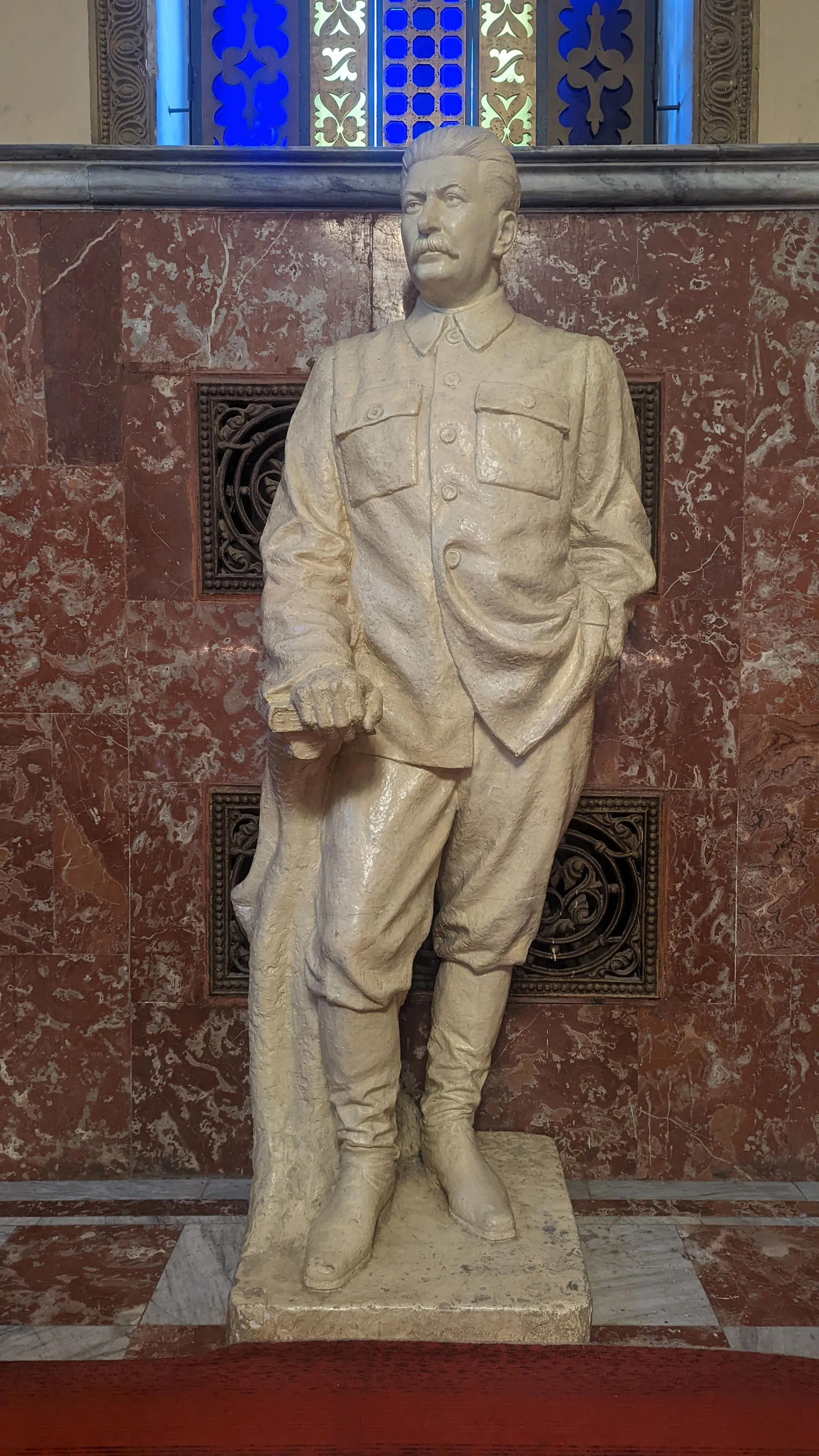 White statue of Joseph Stalin inside the Stalin Museum in Gori, Georgia, against a marble and stained glass backdrop.