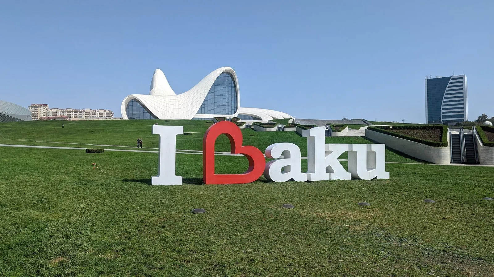 I love Baku sign in front of the Heydar Aliyev Center, with lush green grass and modern buildings in the background.