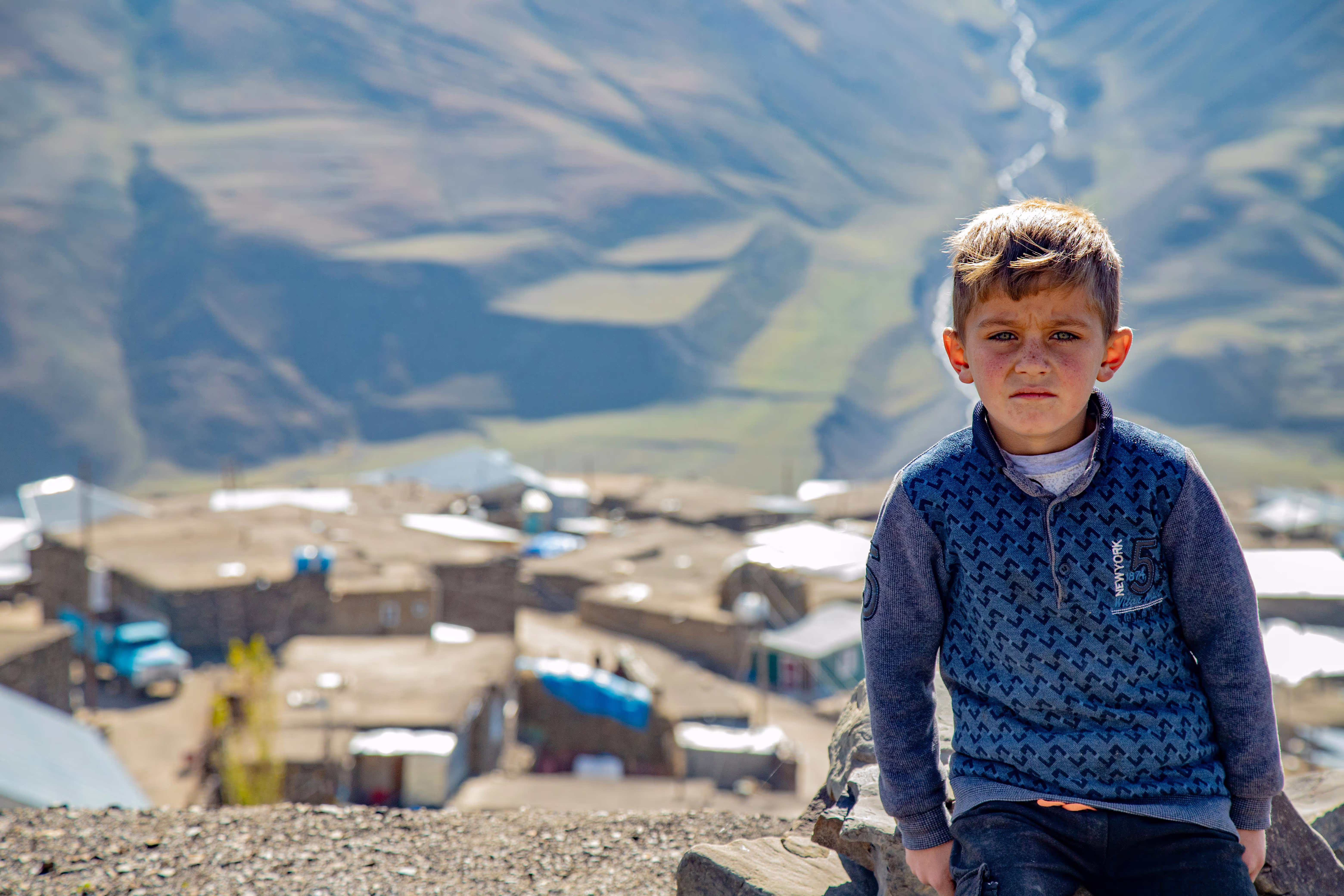 A young boy in Khinaliq, Azerbaijan, sitting on a rock with a stunning mountain village in the background.