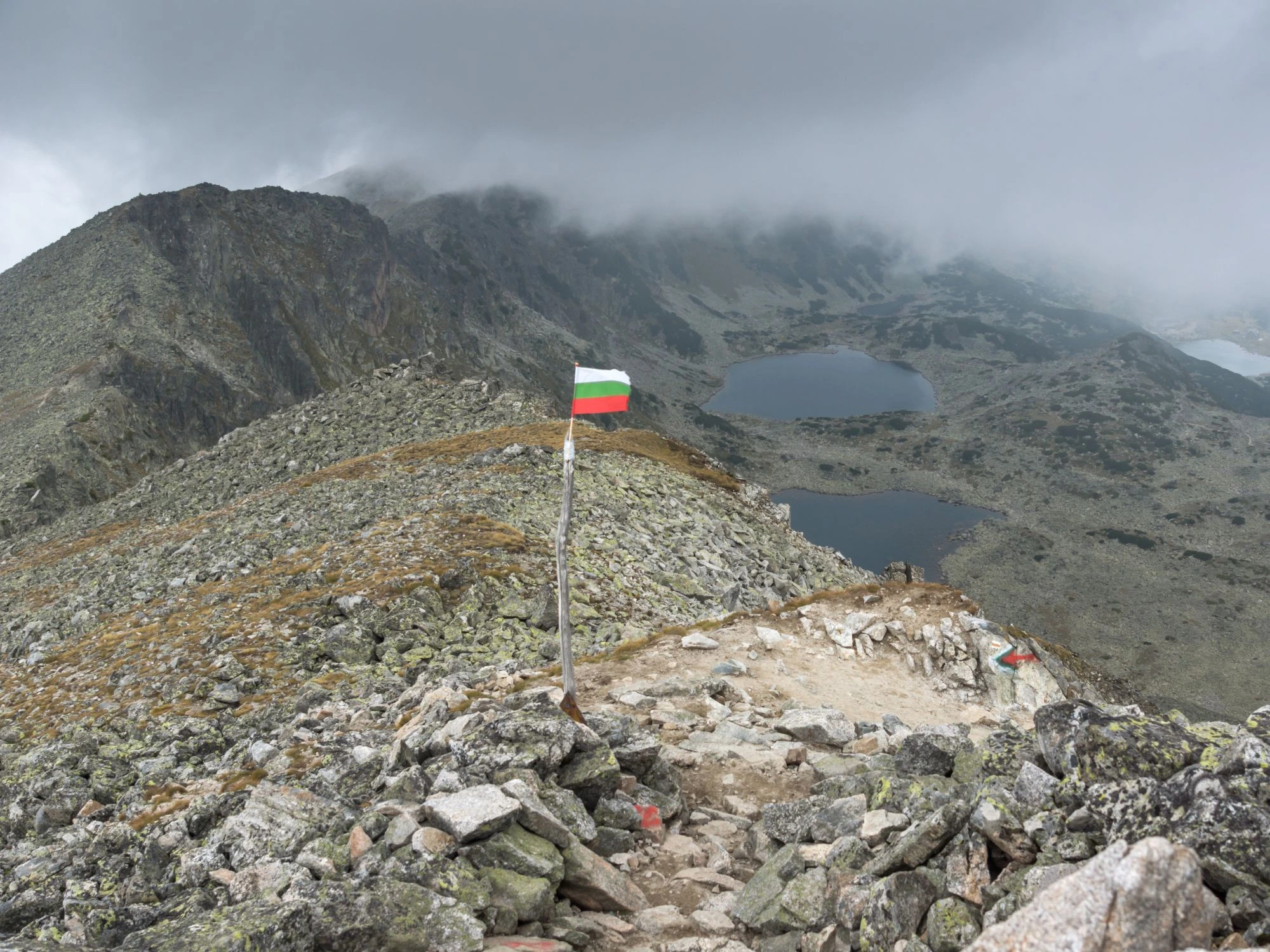 Overcast view from the summit of Musala Peak, Bulgaria, a Bulgarian flag standing against a misty mountain backdrop.