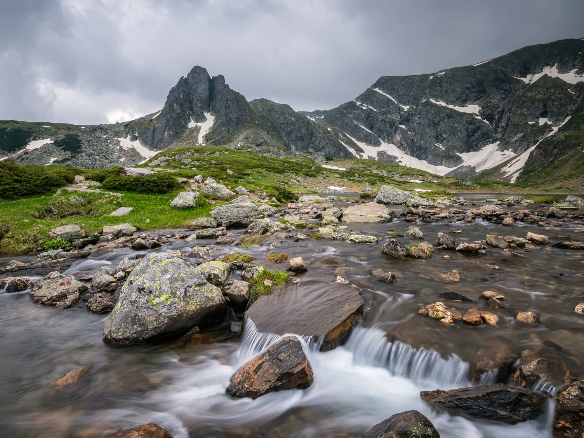 A mountain stream flows over rocks in Rila, Bulgaria, with jagged peaks and patches of snow in the distance.