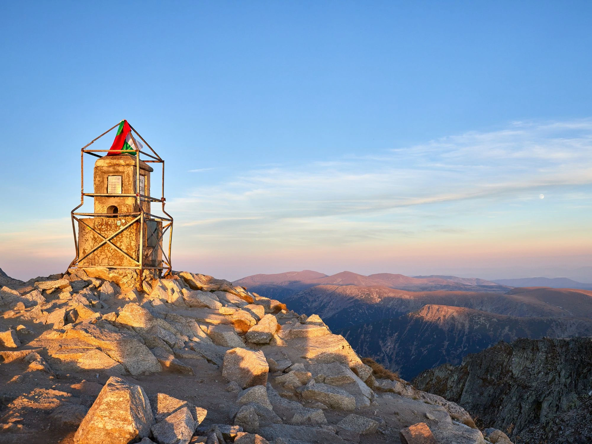 Sunset at Musala Peak, Bulgaria, with golden light illuminating the rugged summit and distant mountain ridges.