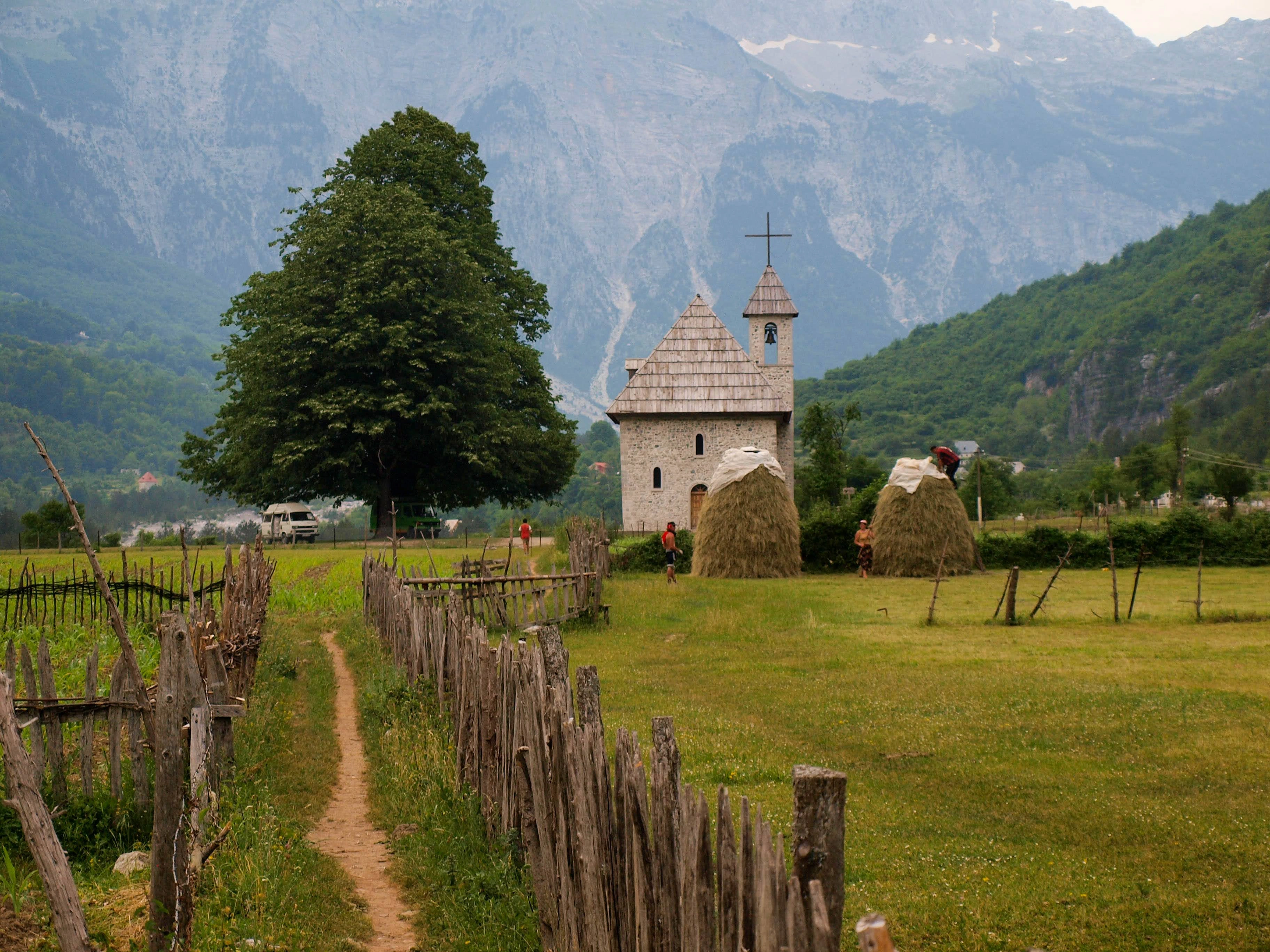 A rustic stone church with a wooden roof, surrounded by haystacks and mountains in Theth, Albania.