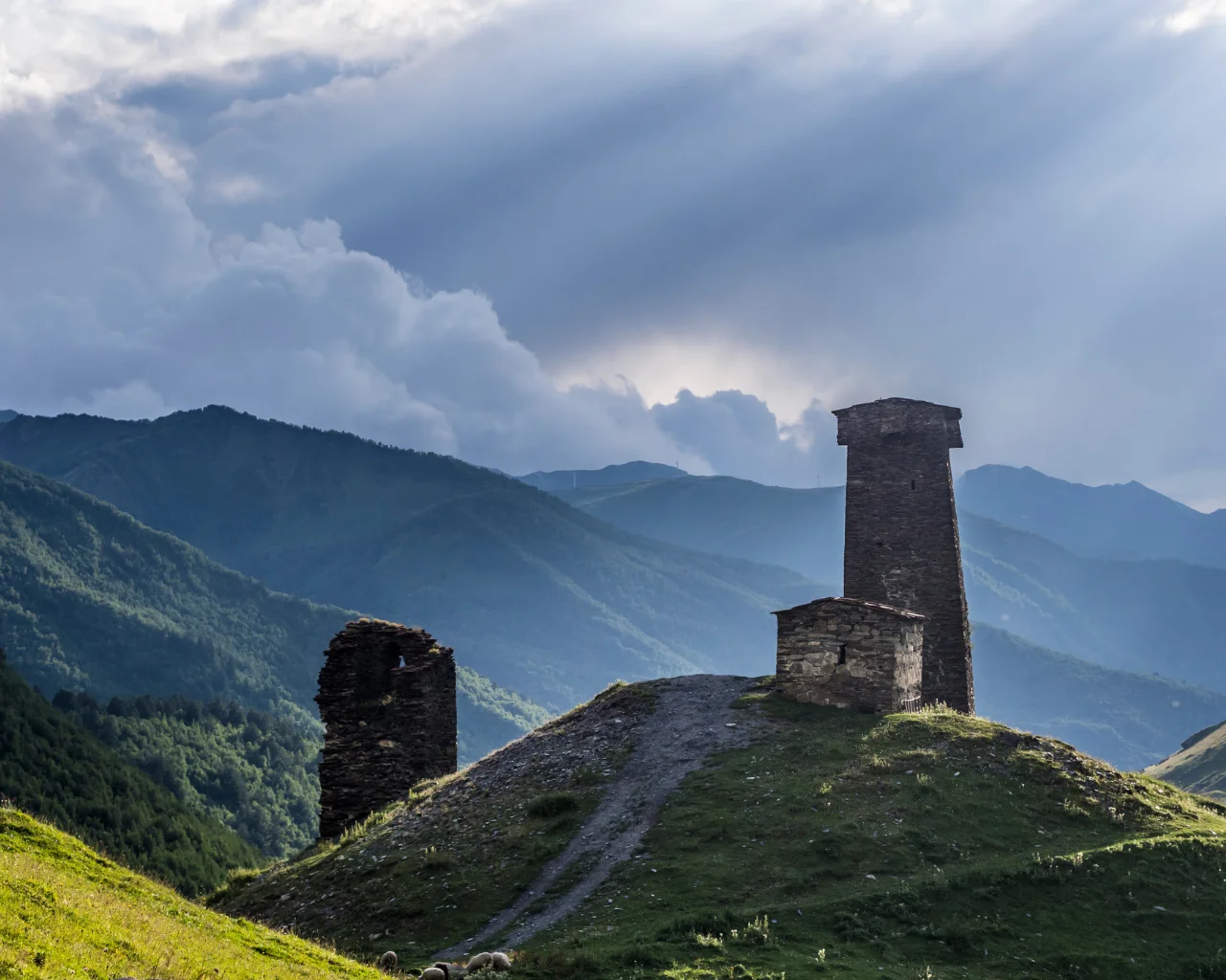 Ancient stone towers on a grassy hill in Svaneti, Georgia, with forested mountains behind.