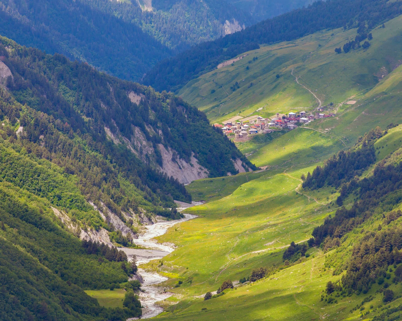 A small Georgian village with red-roofed houses is nestled among lush green hills and a winding river in Svaneti.