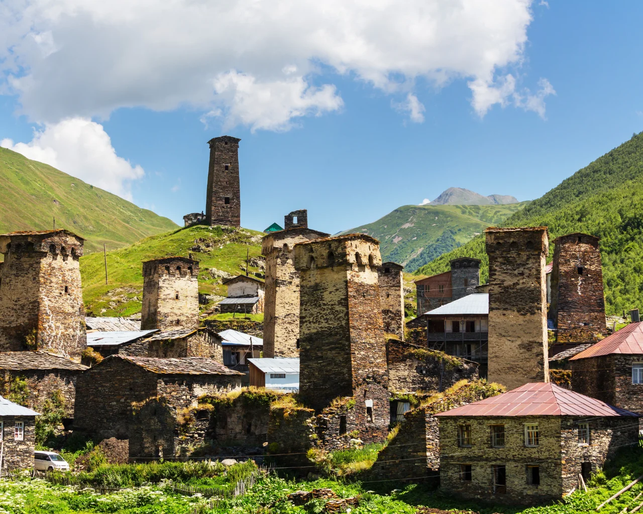 Stone defensive towers dominate the mountain village of Ushguli, Georgia, under a bright blue sky with scattered clouds.