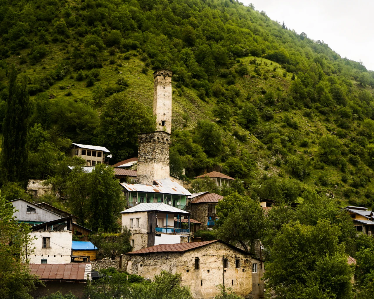 A historic stone tower rises among rustic houses in a green hillside village in Svaneti, Georgia, surrounded by trees.