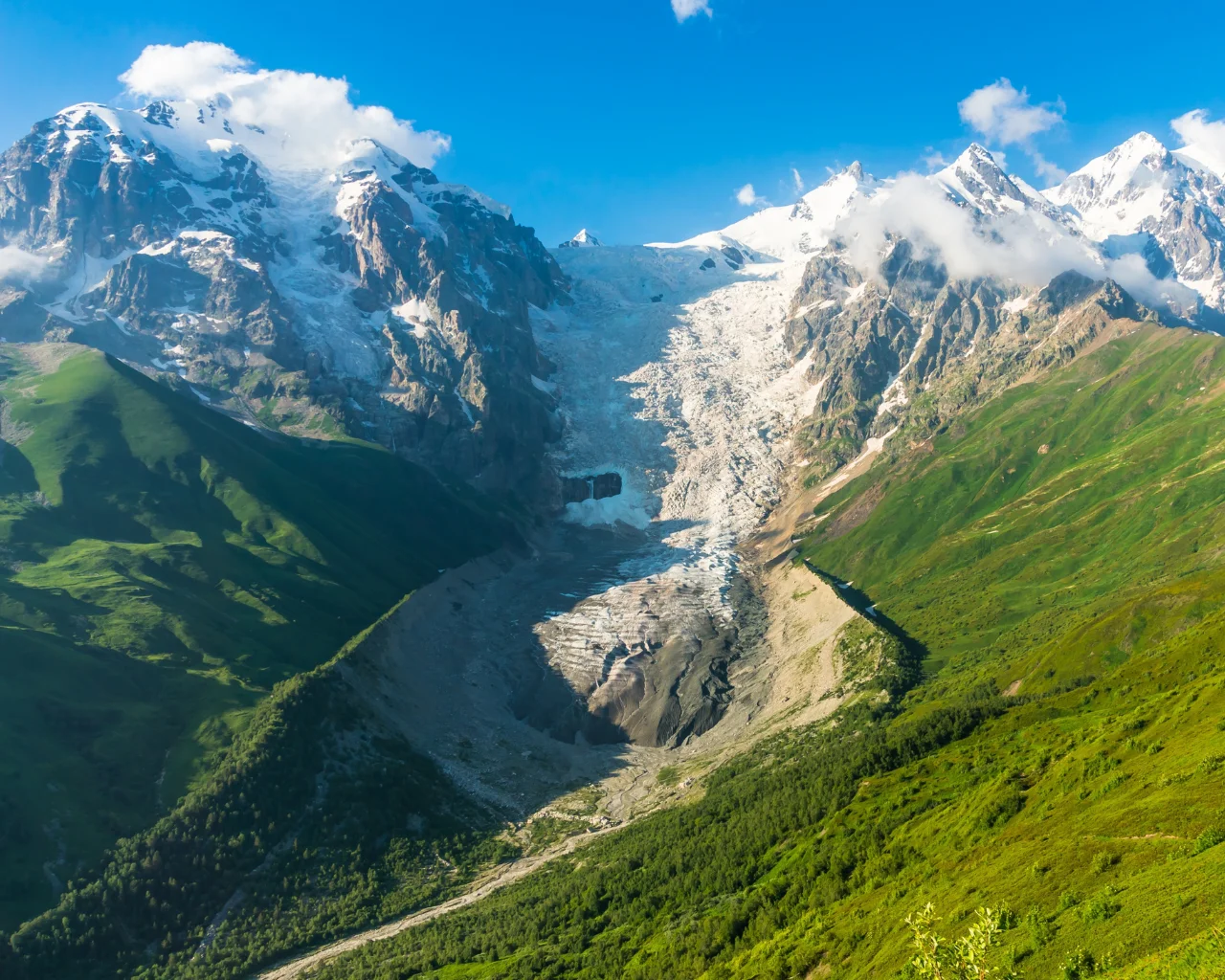  A stunning view of Kazbek Glacier cascading between rugged, snow-capped peaks above a lush green valley.