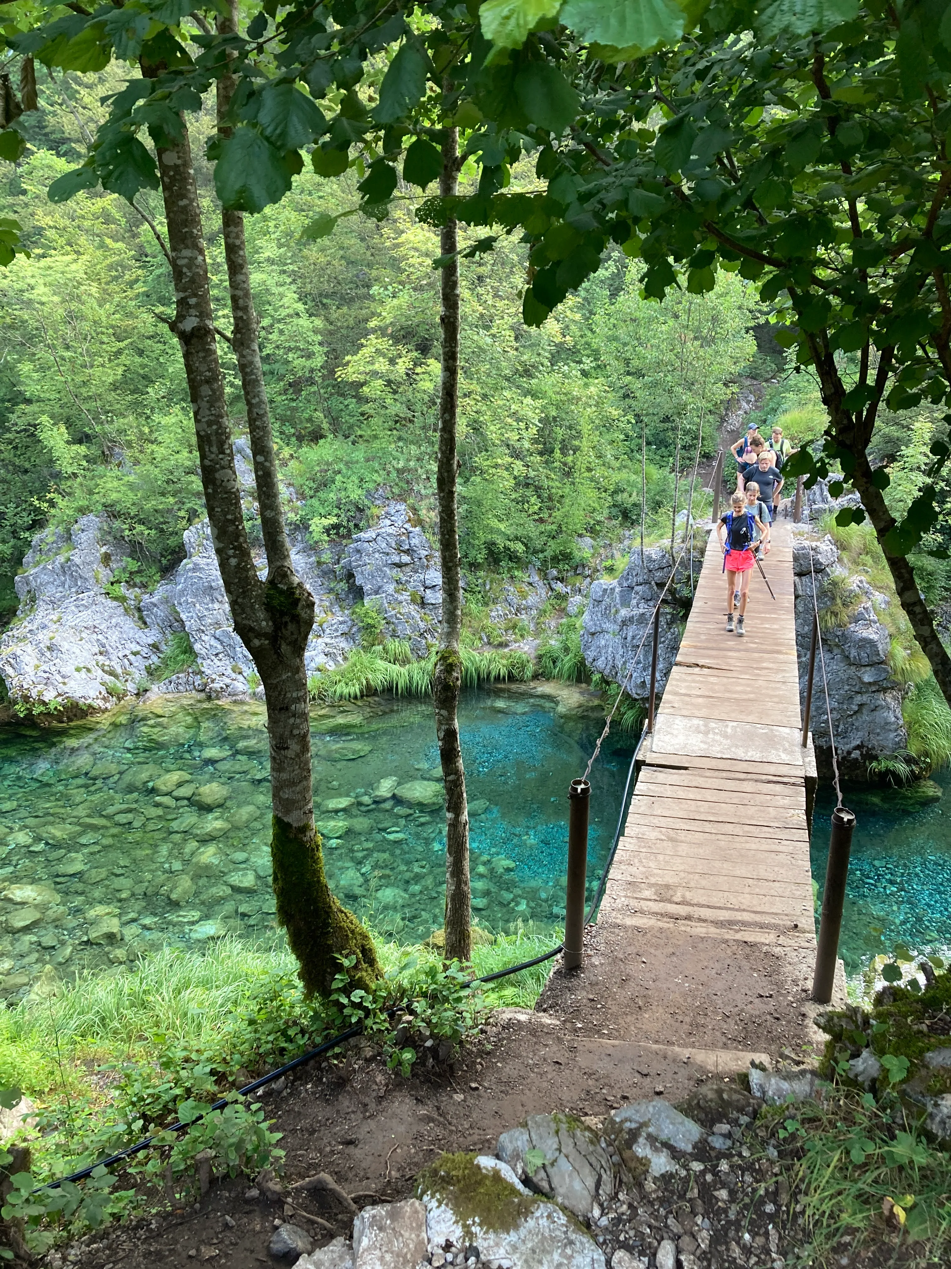 A group of hikers crossing a wooden bridge over a turquoise river in Theth, Albania, surrounded by lush green forests