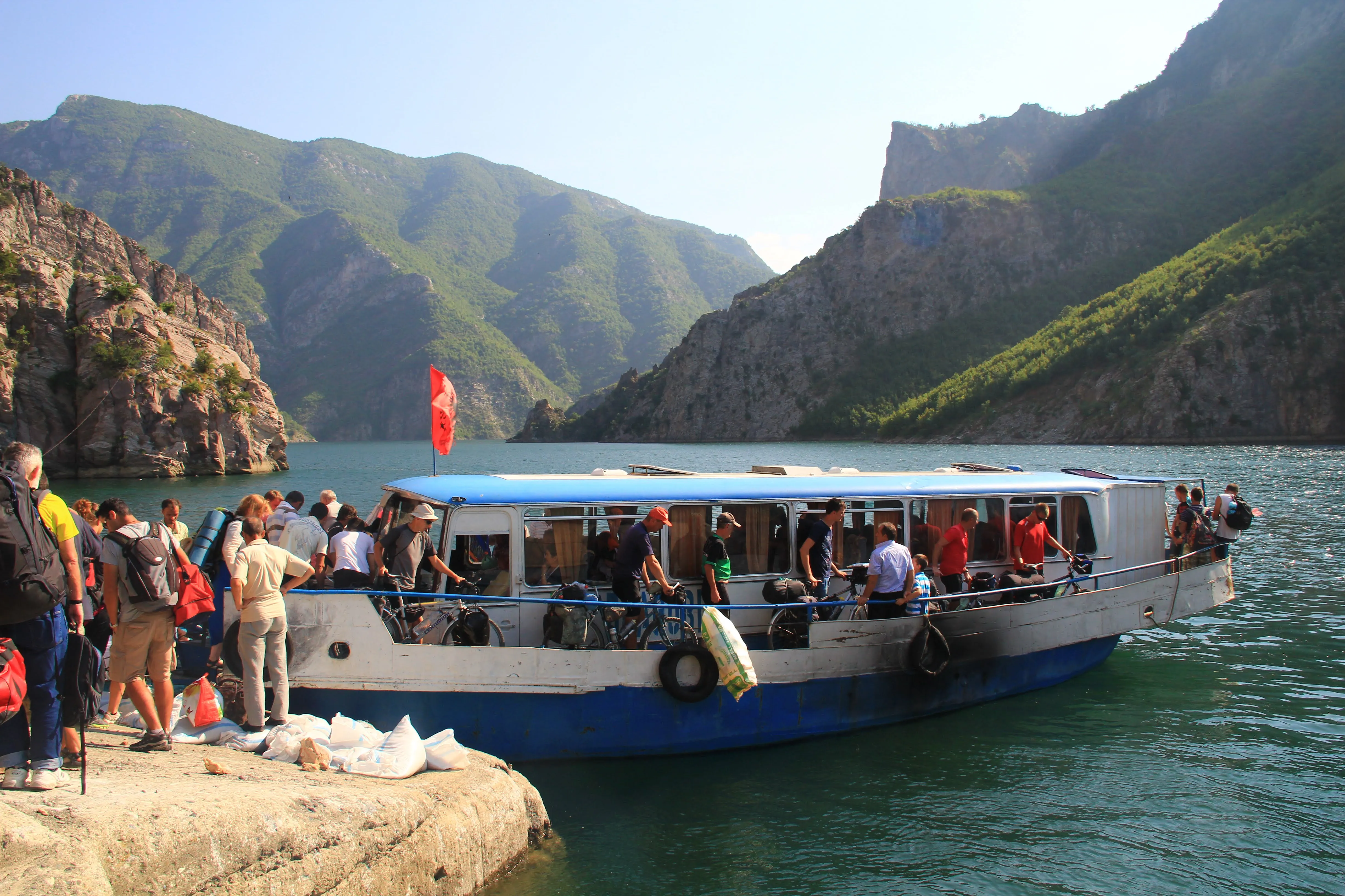 Travelers boarding a small ferry on Koman Lake, Albania, with green mountains and dramatic rocky cliffs in the background.