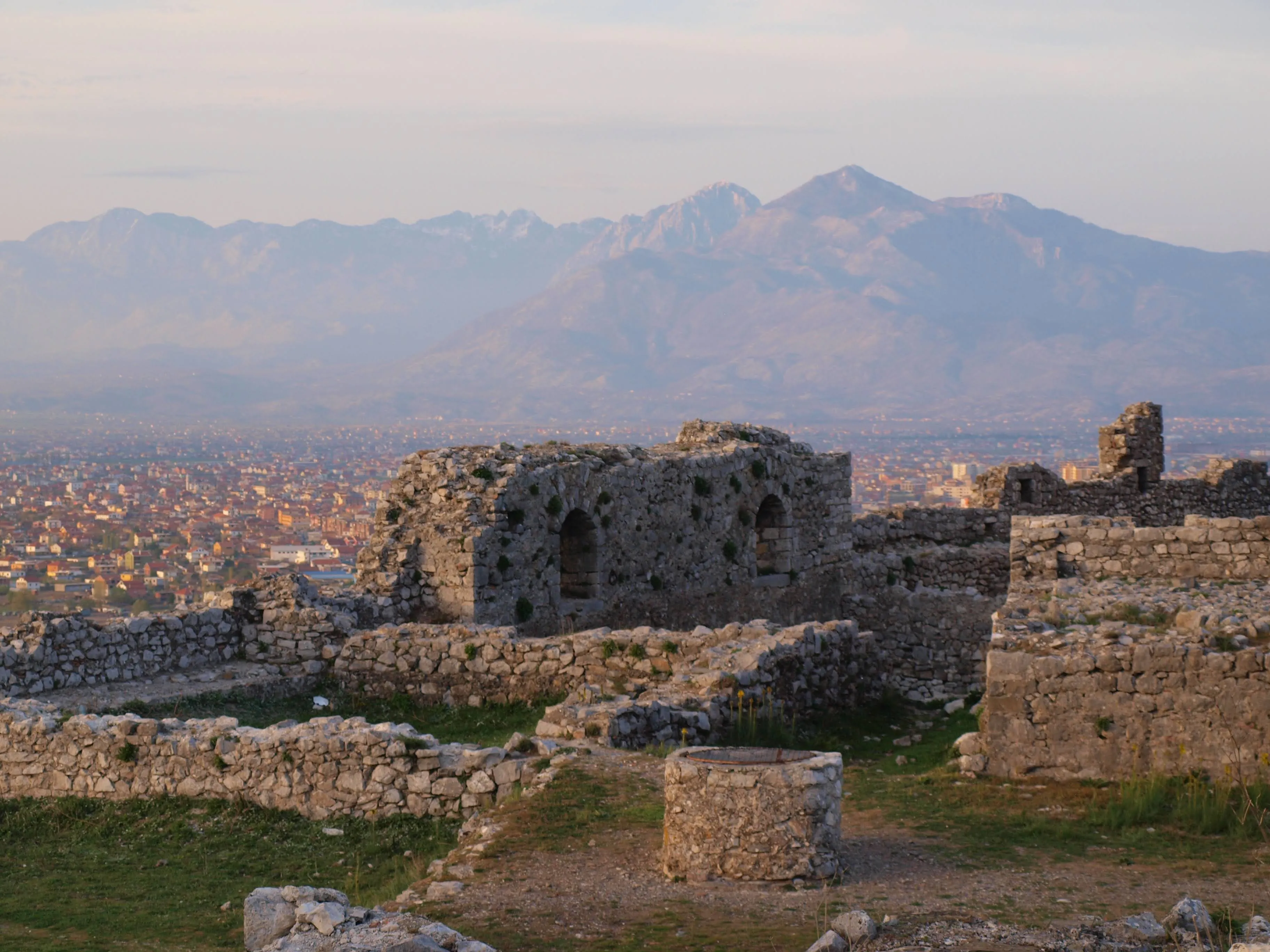 The ruins of Rozafa Castle overlooking Shkodra, Albania, with distant mountain peaks under a golden sunset glow.