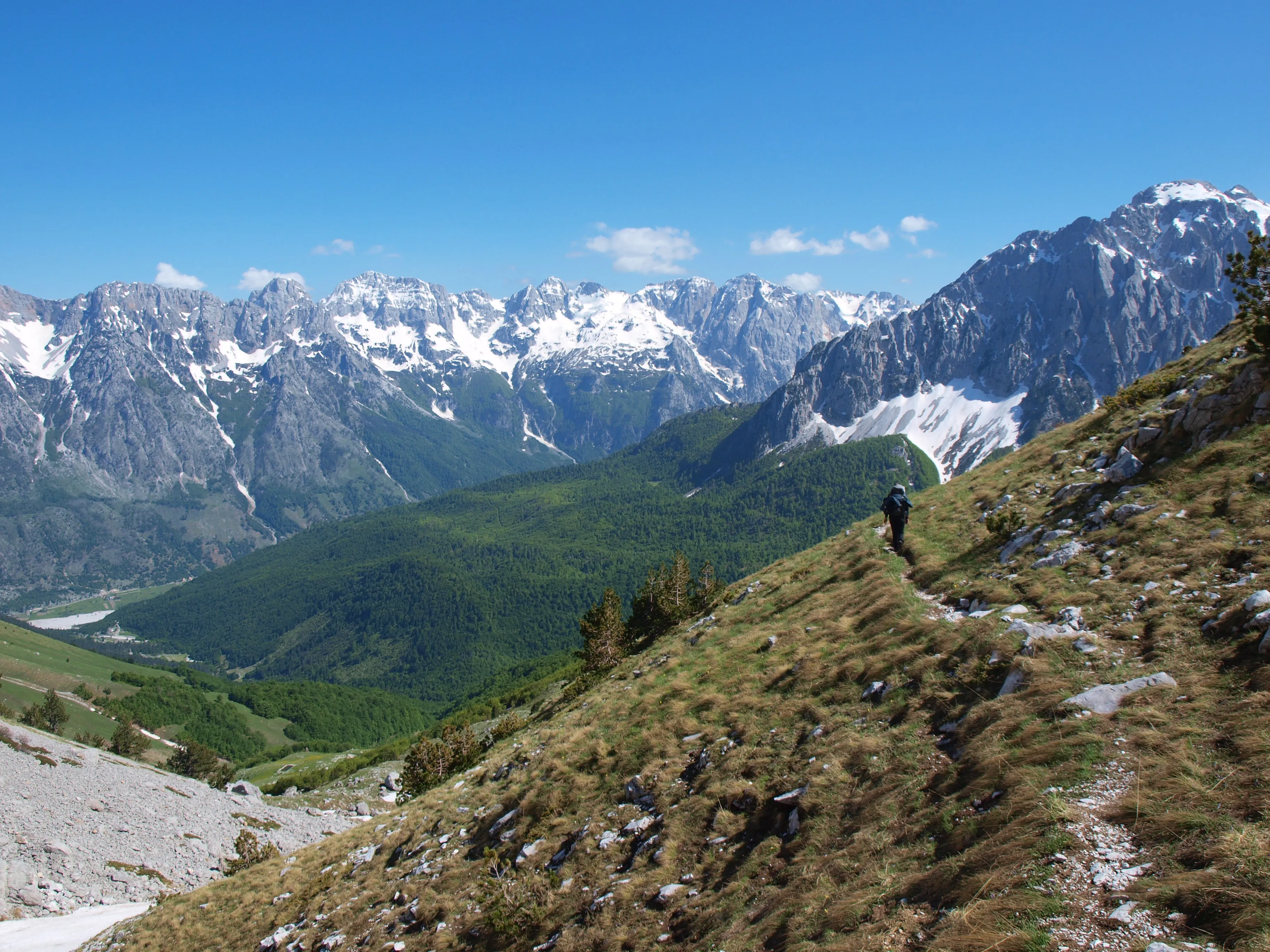 Scenic hiking trail in Valbona Valley, Albania, with a lone hiker traversing steep alpine slopes toward snow-capped peaks.