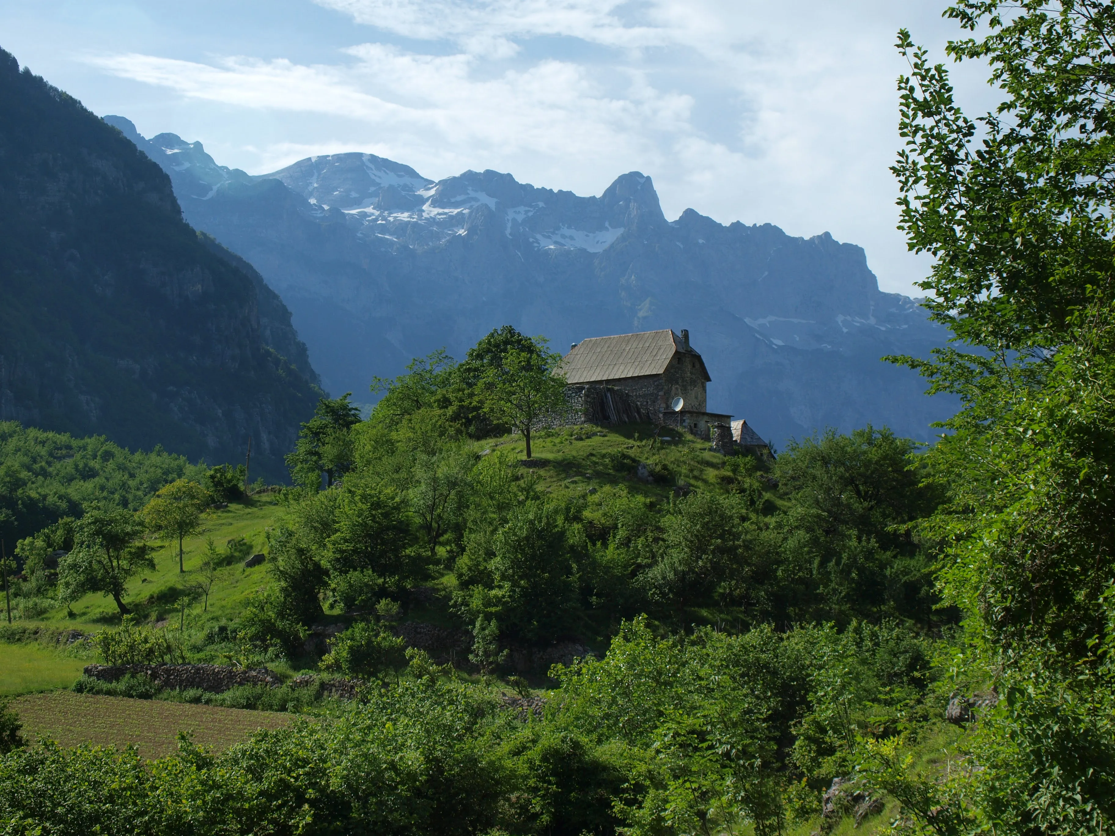 Remote stone house on a lush hill in Theth, Albania, with towering alpine peaks in the background.
