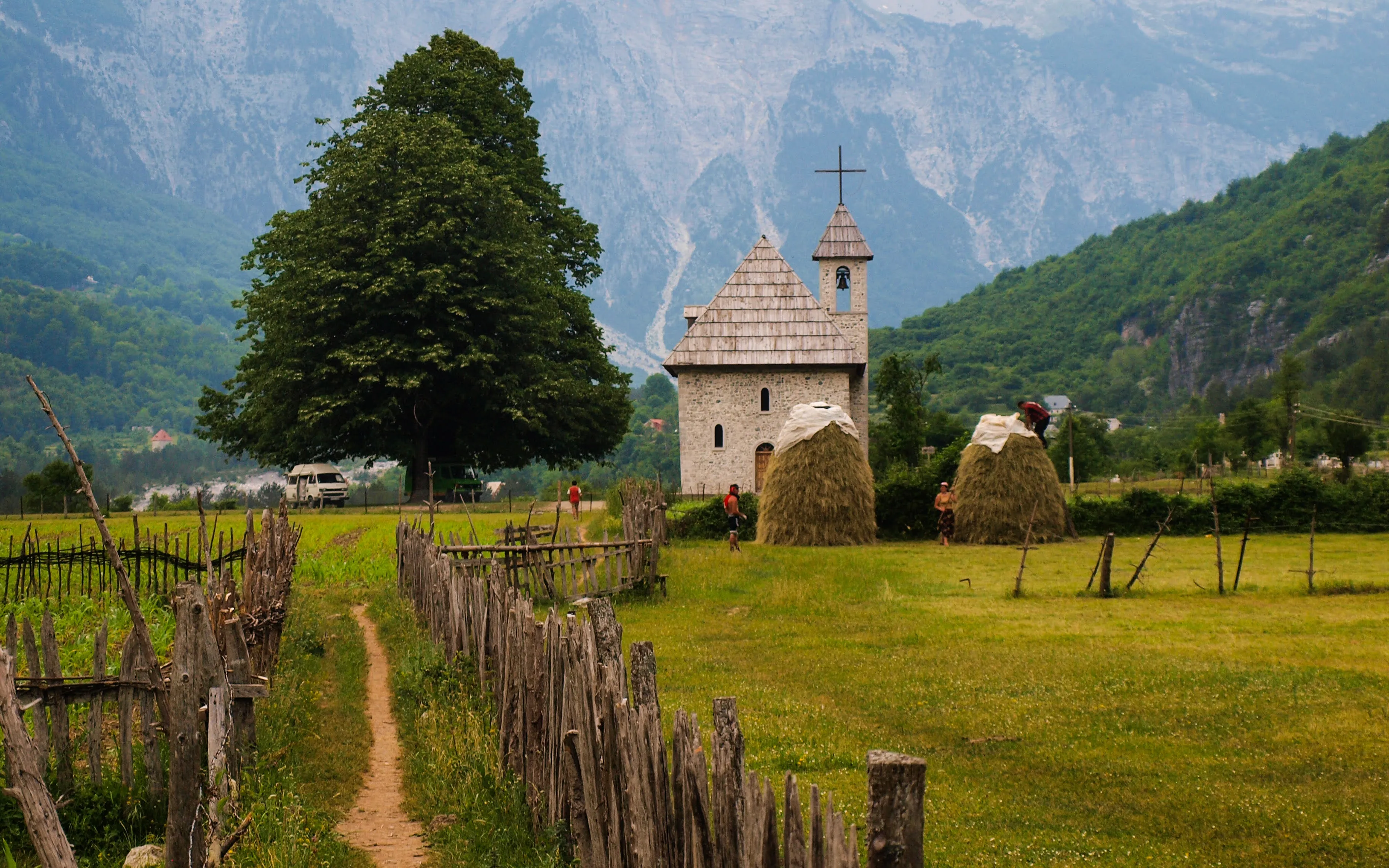 Theth church in Albania, a historic stone building amid green fields, with haystacks and the Accursed Mountains behind.