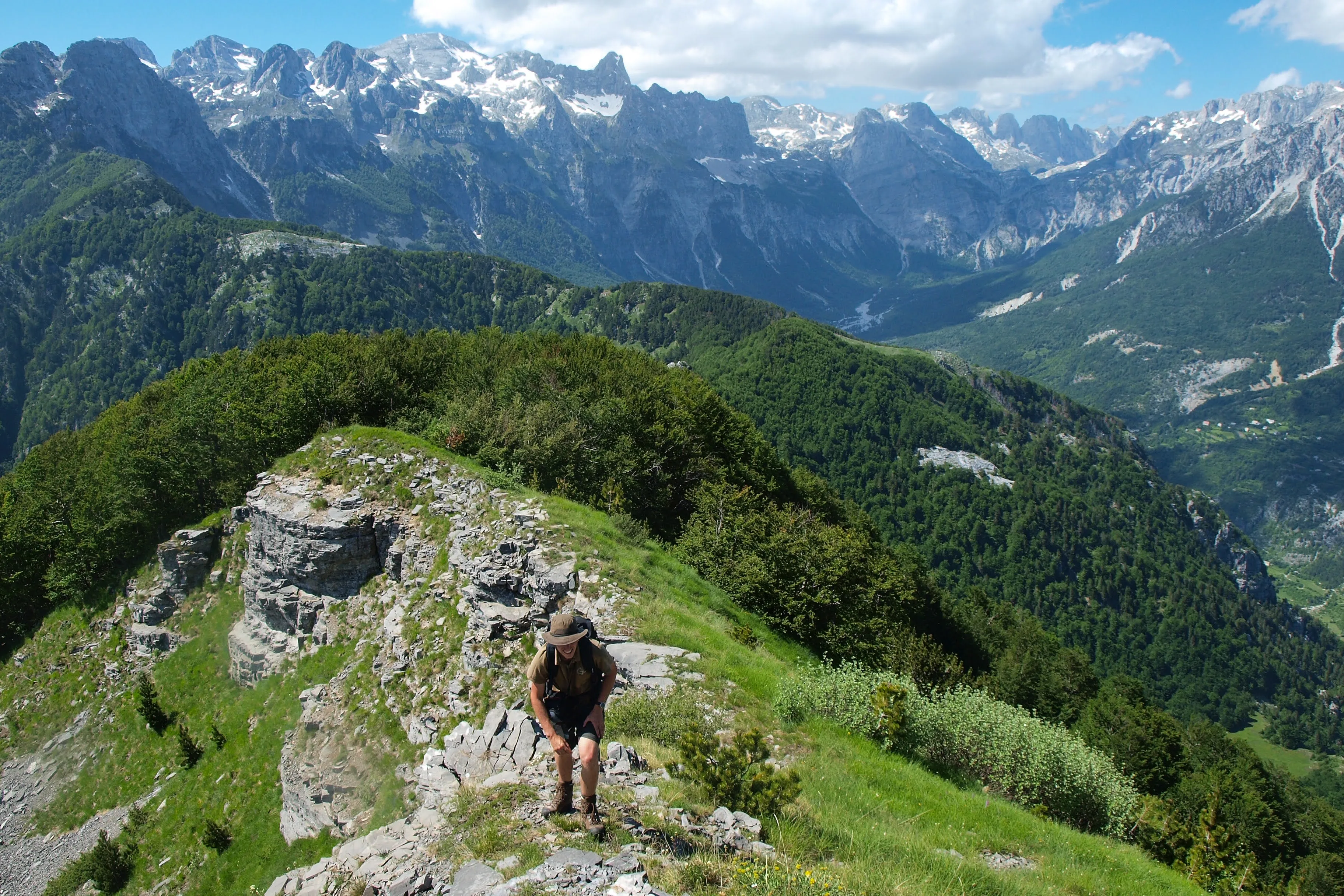 A hiker trekking along a rocky ridge in the Albanian Alps, with panoramic views of snow-capped peaks and deep valleys
