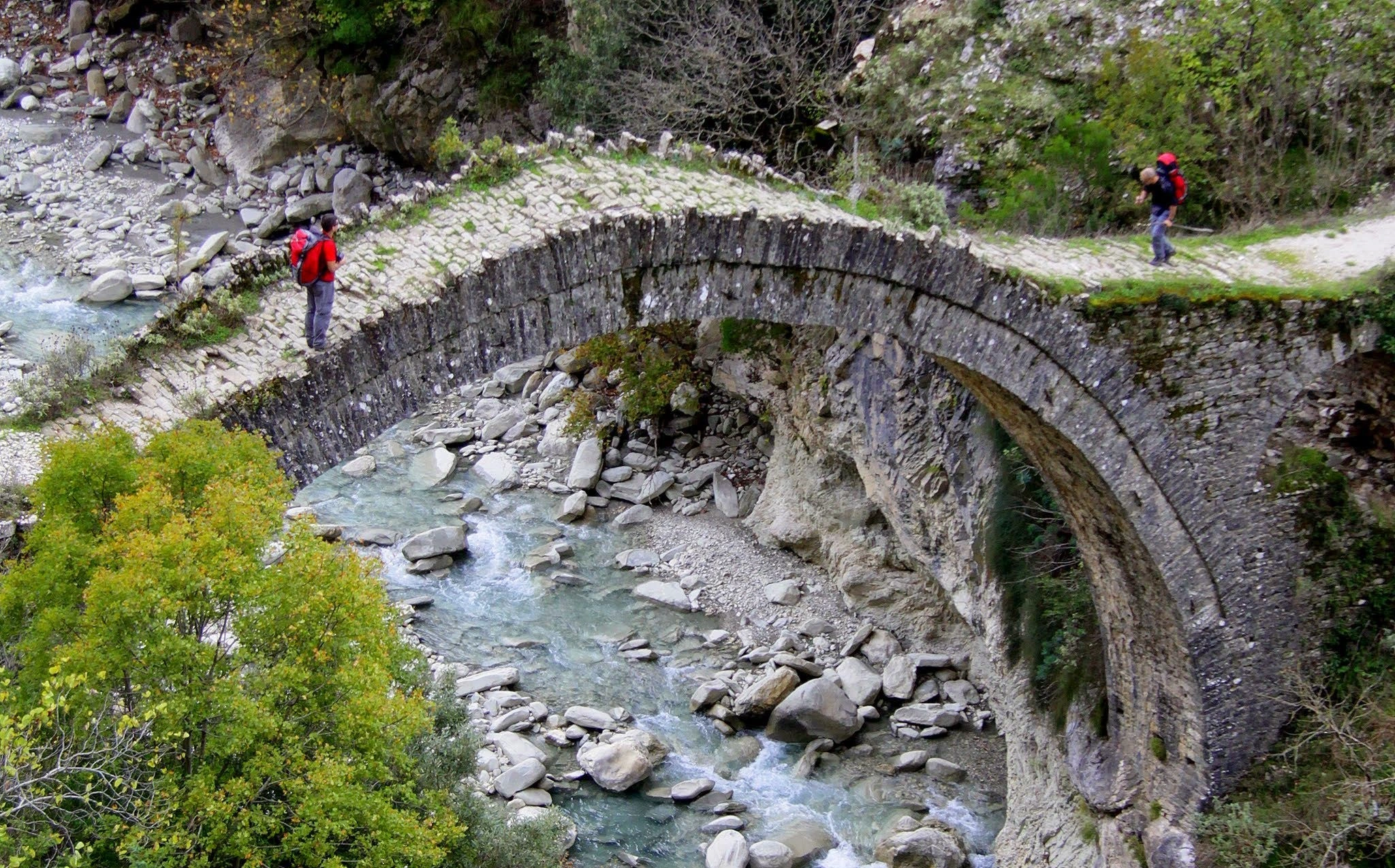 A historic stone bridge arching over a rocky river, with hikers crossing and lush trees in the background.