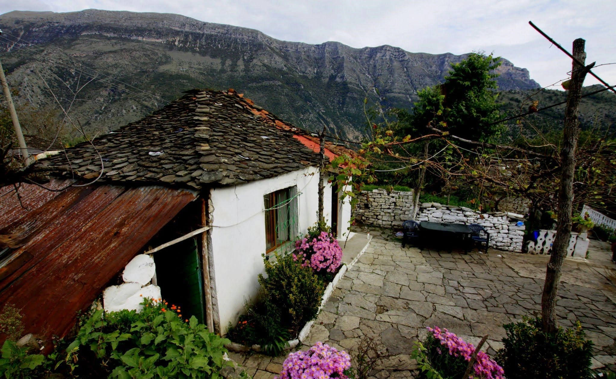 A rustic courtyard with stone pathways, pink flowers, and a small white house against a mountain backdrop.