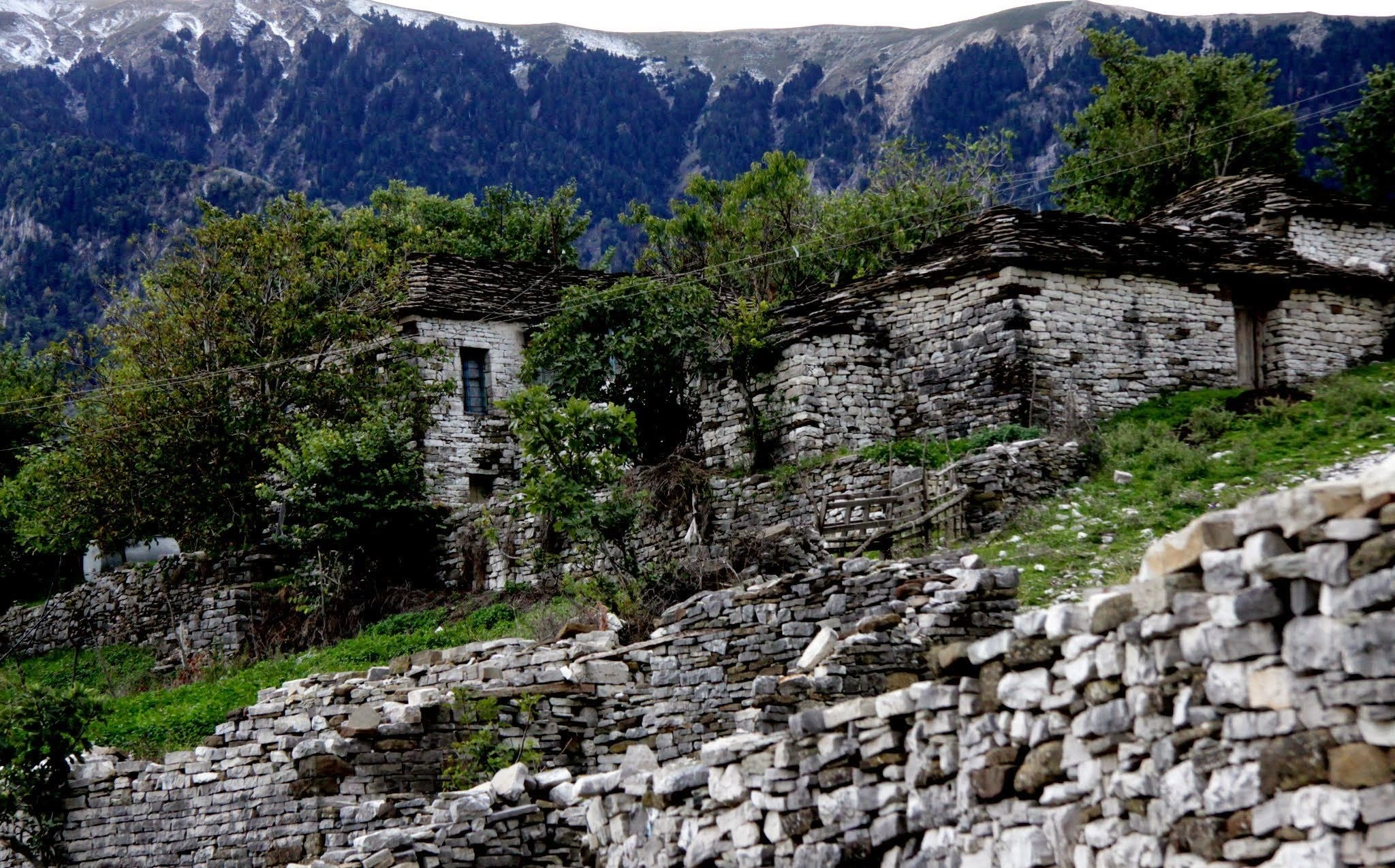 Stone houses in a remote mountain village, surrounded by lush greenery and towering peaks in the background.