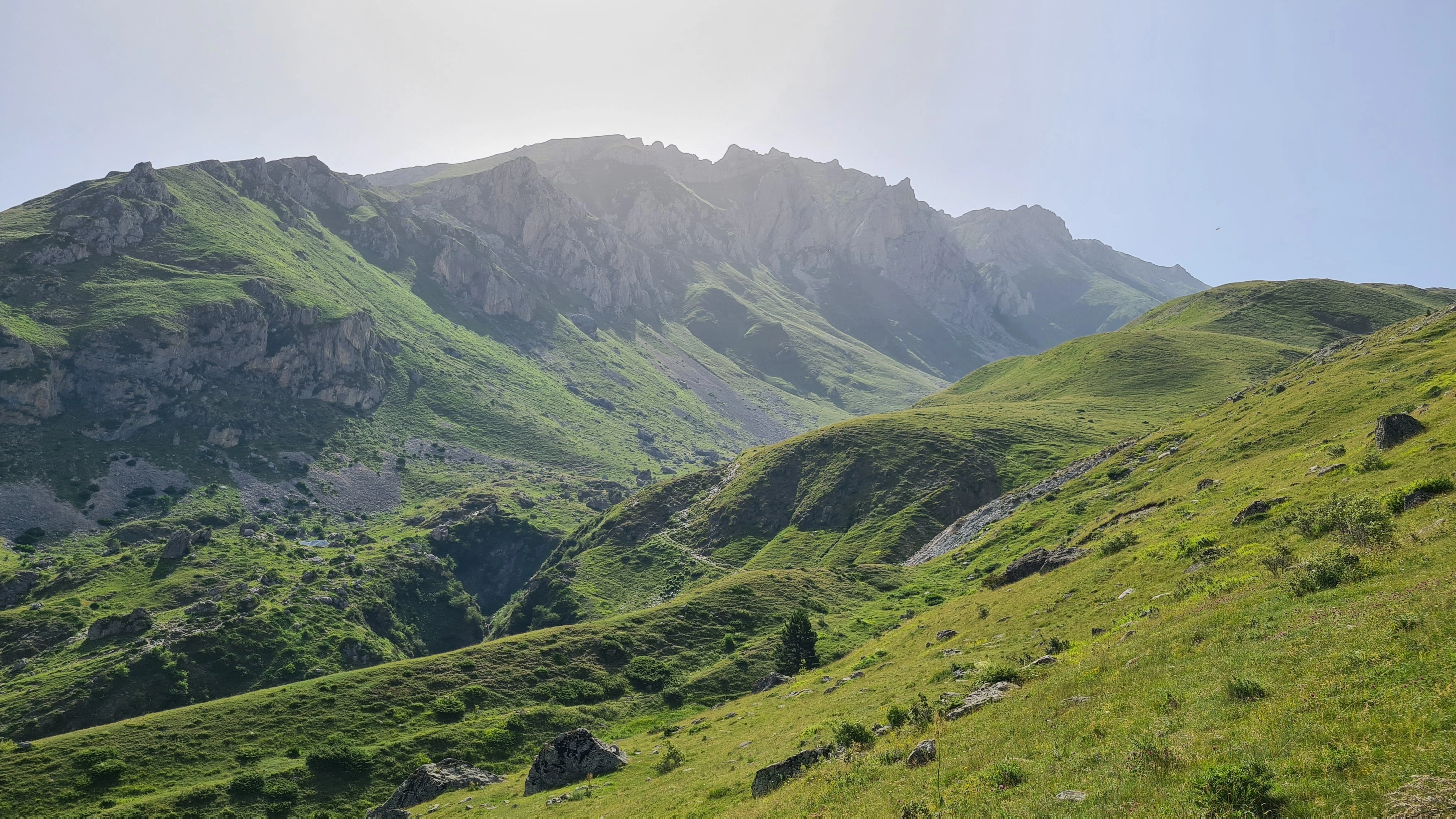 Rolling green hills and towering rocky peaks stretch under bright sunlight in High Scardus, Albania.
