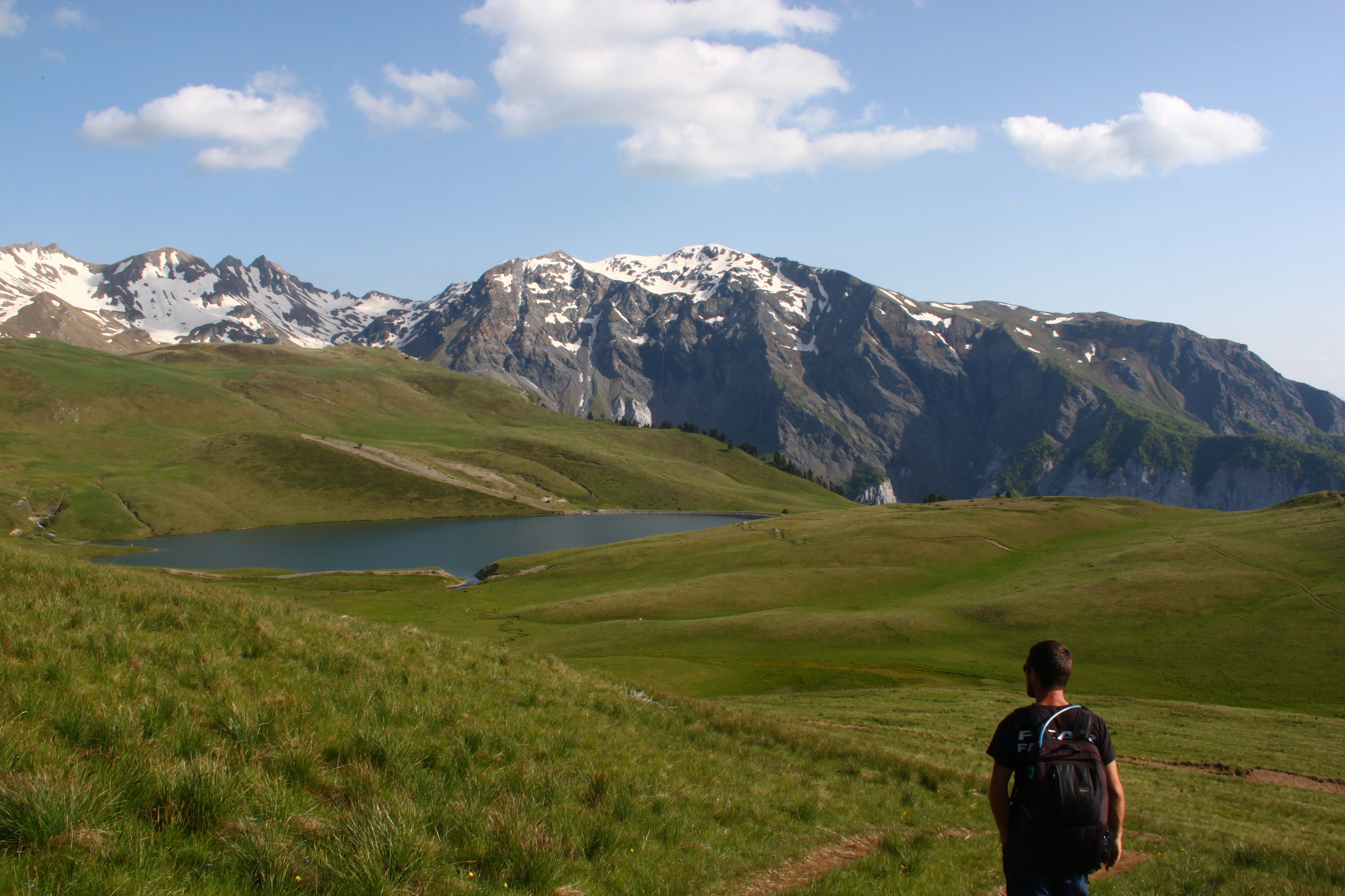 Hiker at glacial lake – A lone hiker with a backpack stands near a blue glacial lake, with snowy peaks in the distance.