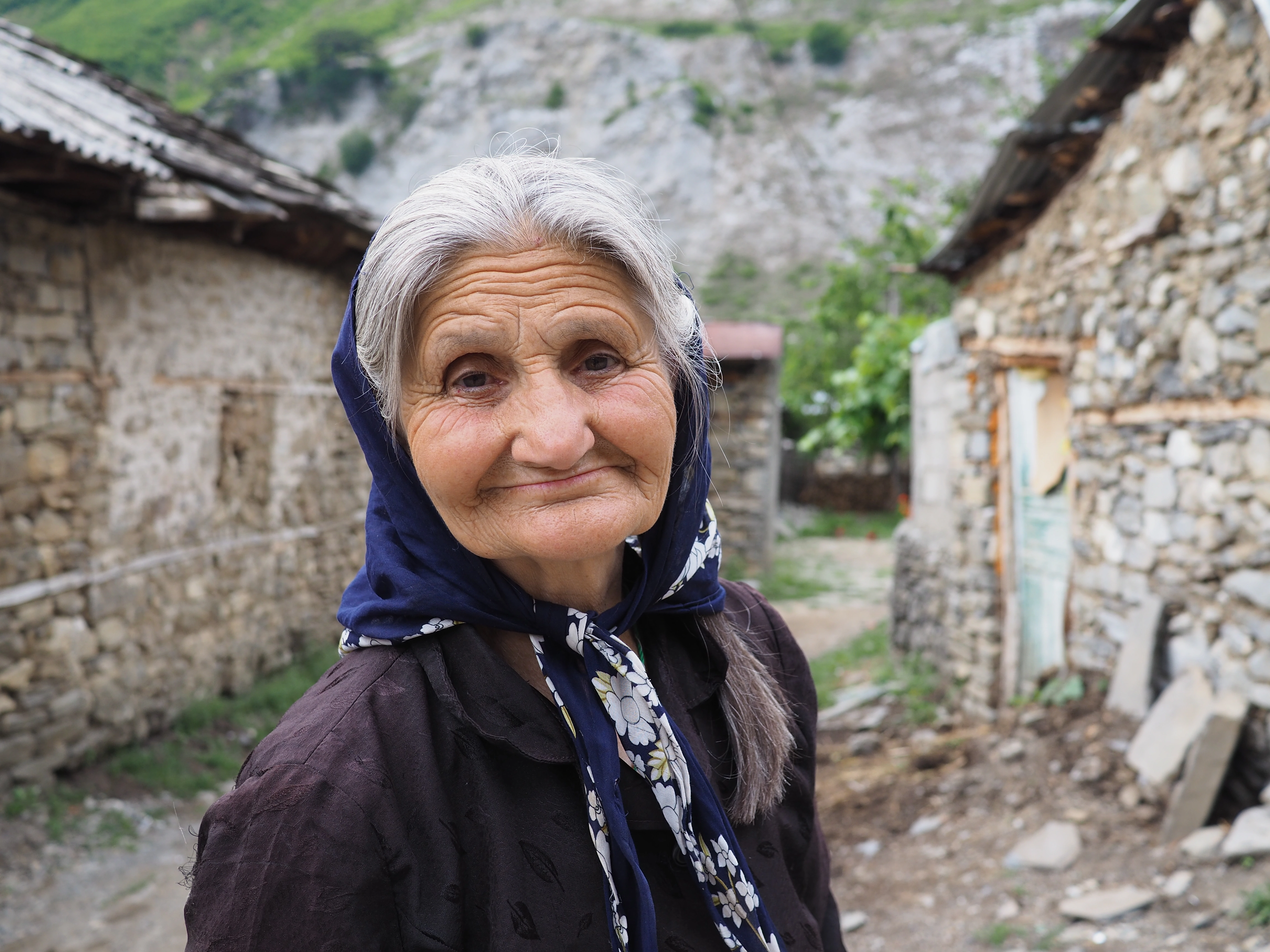 A smiling elderly woman in a blue headscarf stands between rustic stone houses in a mountain village.