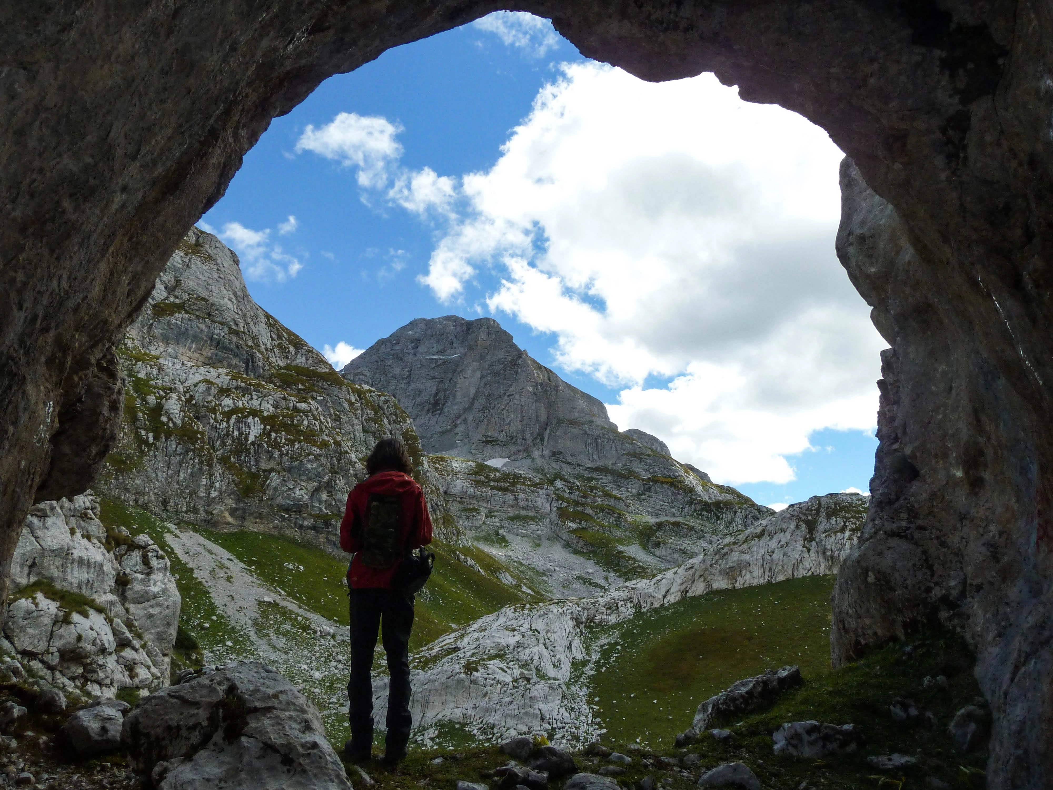 A traveler standing inside a cave, gazing at the mountain range under a blue sky.