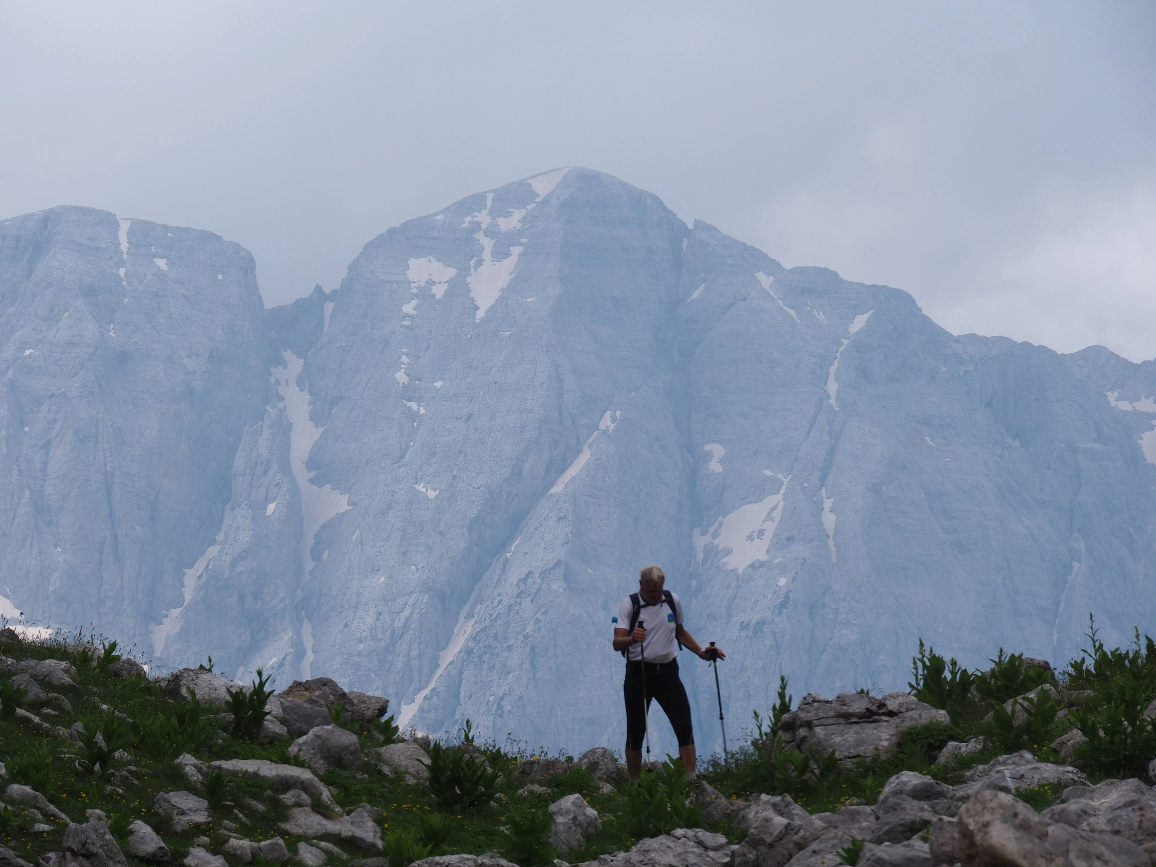 A lone hiker trekking through rocky terrain with towering, snow-dusted peaks in the distance.