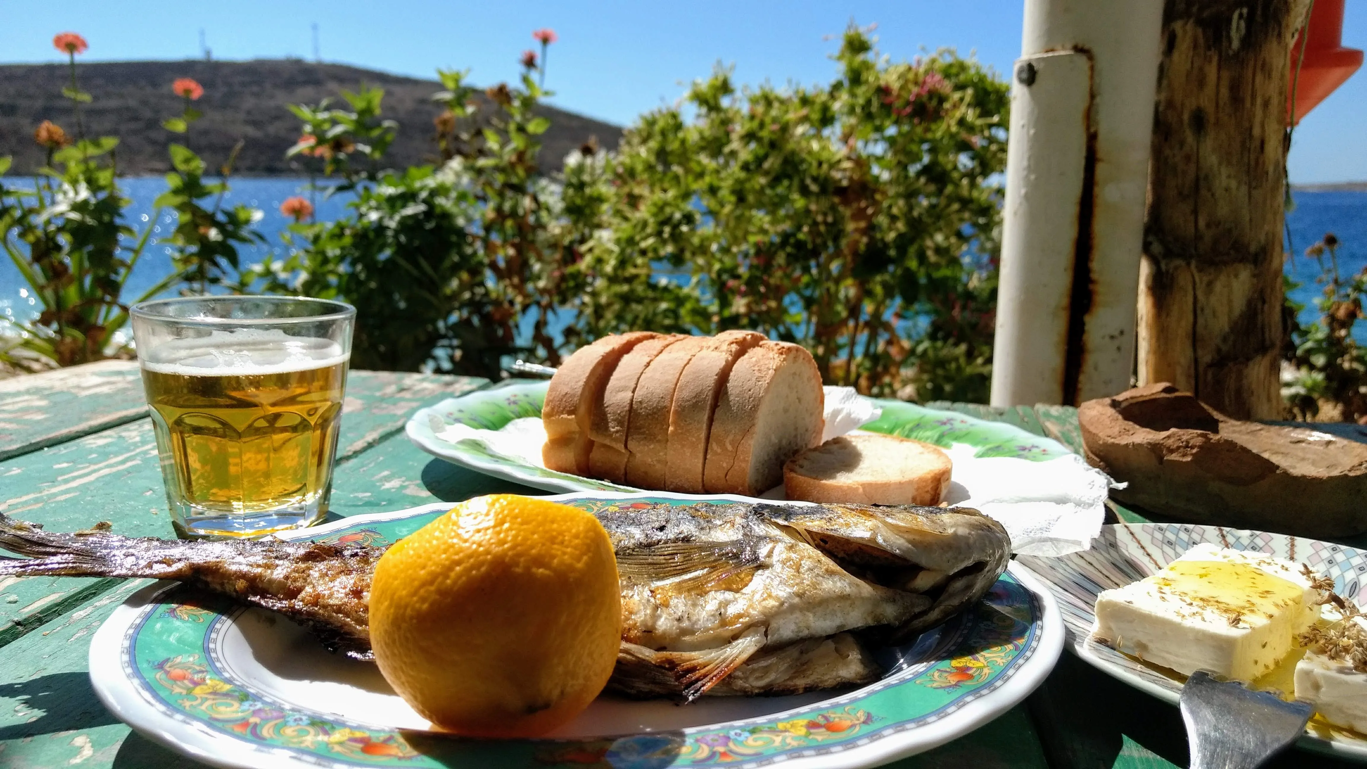 Freshly grilled fish with lemon, feta cheese, and bread, served with a glass of beer at a seaside restaurant.