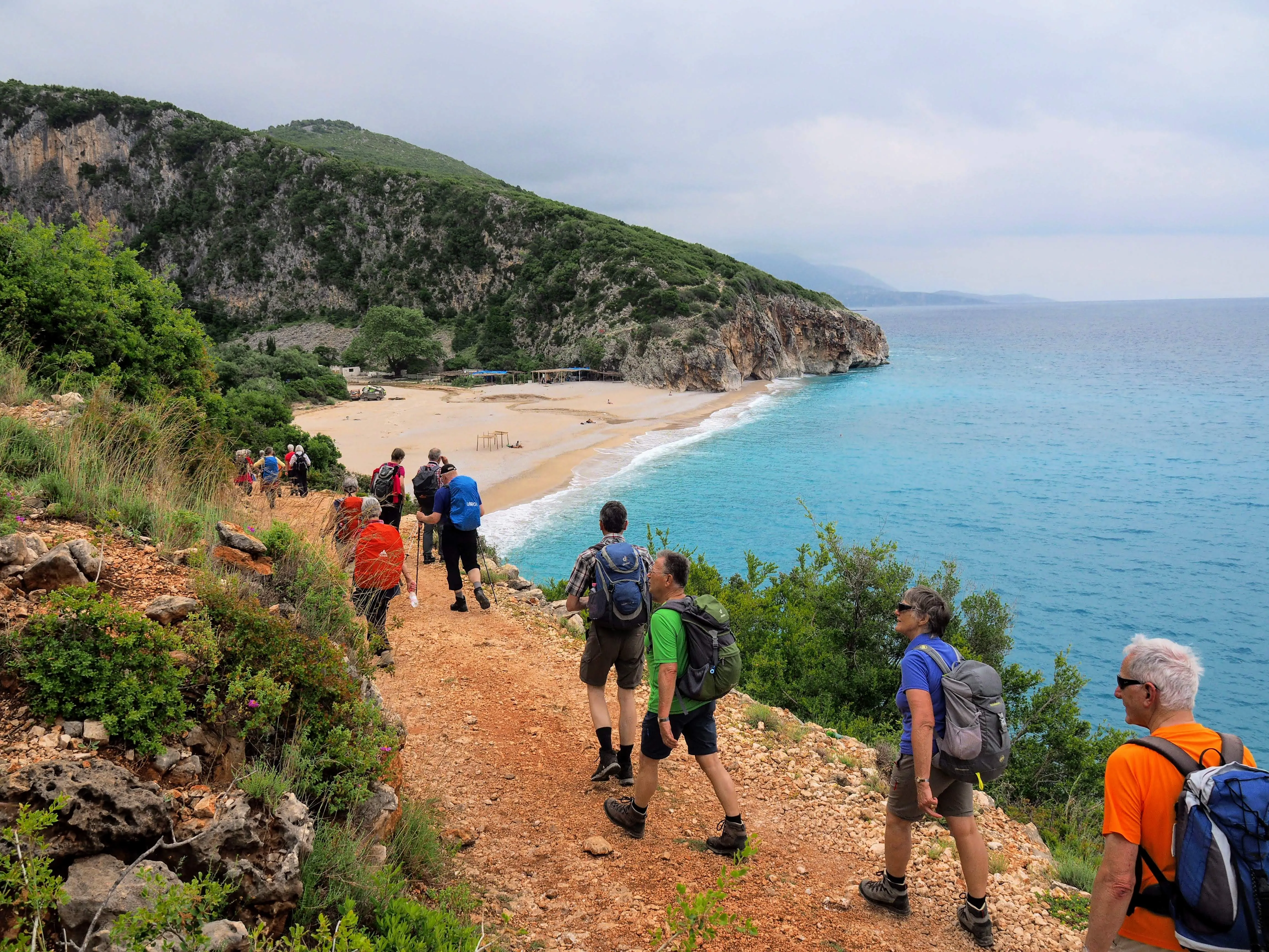 Hikers trekking down a rugged trail towards a secluded sandy beach with turquoise waters along the Albanian coast.