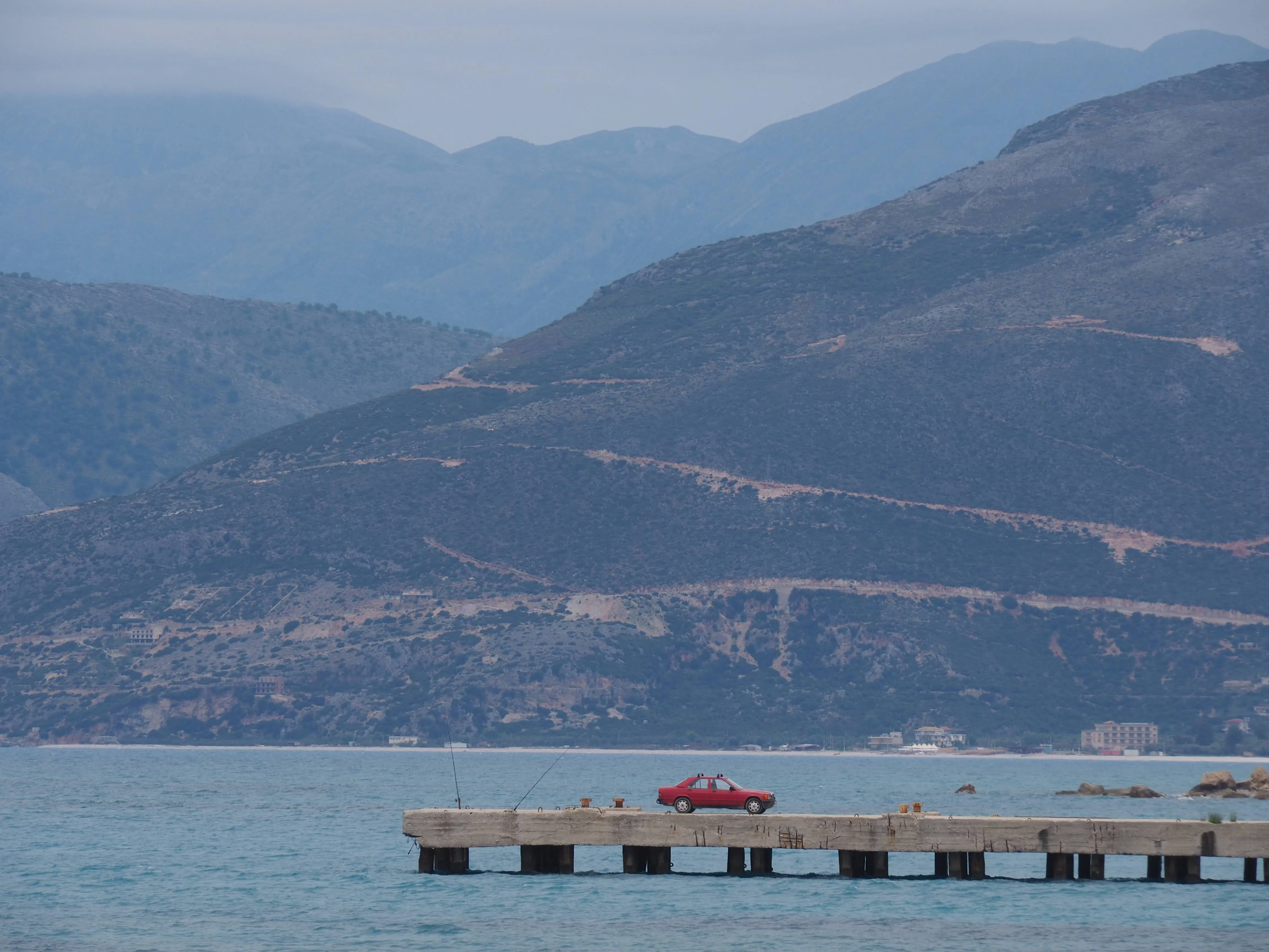 A red car parked at the edge of a pier with rugged mountains in the background along the Albanian Riviera.