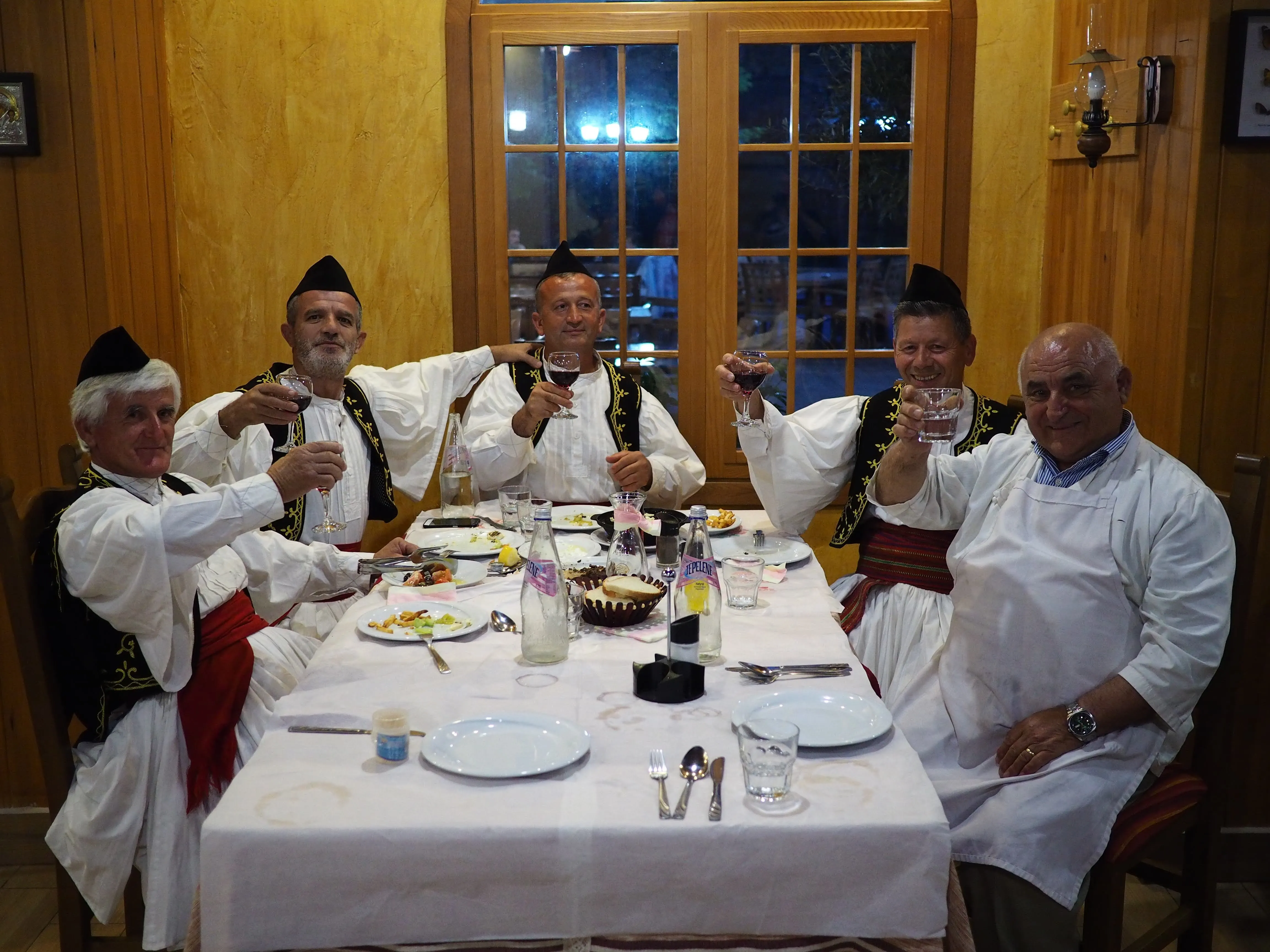 A group of men in traditional Albanian clothing raising glasses in a cozy wooden restaurant, enjoying local cuisine.
