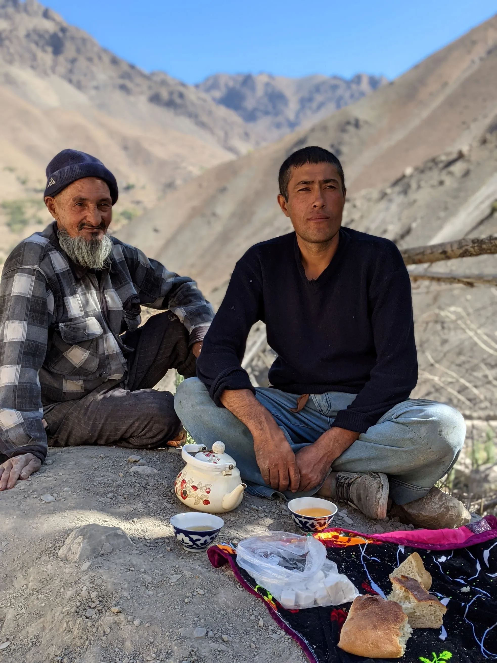 Tajikistan hospitality: Two men enjoy traditional tea and bread in the mountains, showcasing local culture and warmth.