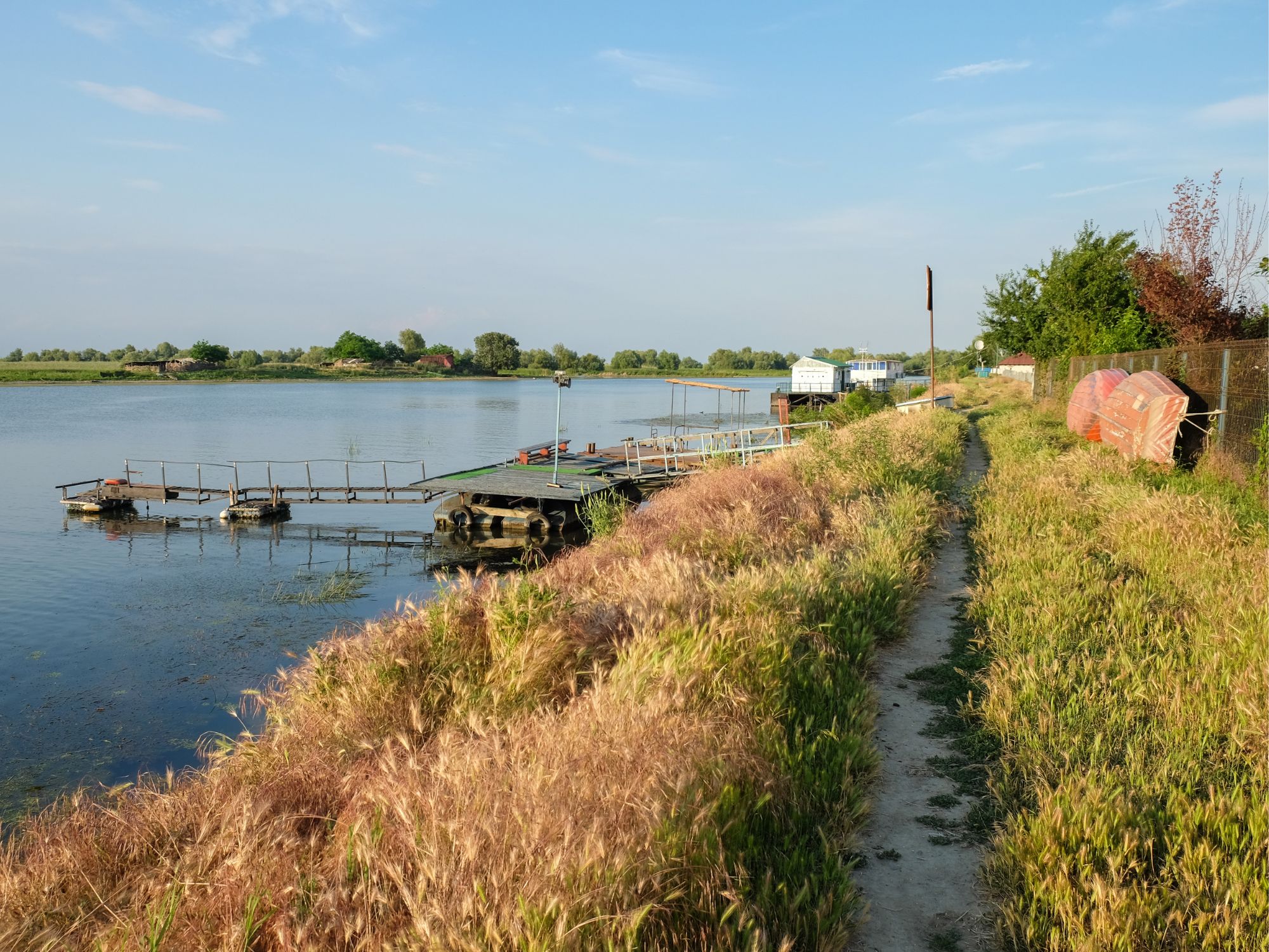 A riverside path lined with tall grass, leading to wooden docks and floating houses in a quiet Danube Delta village.