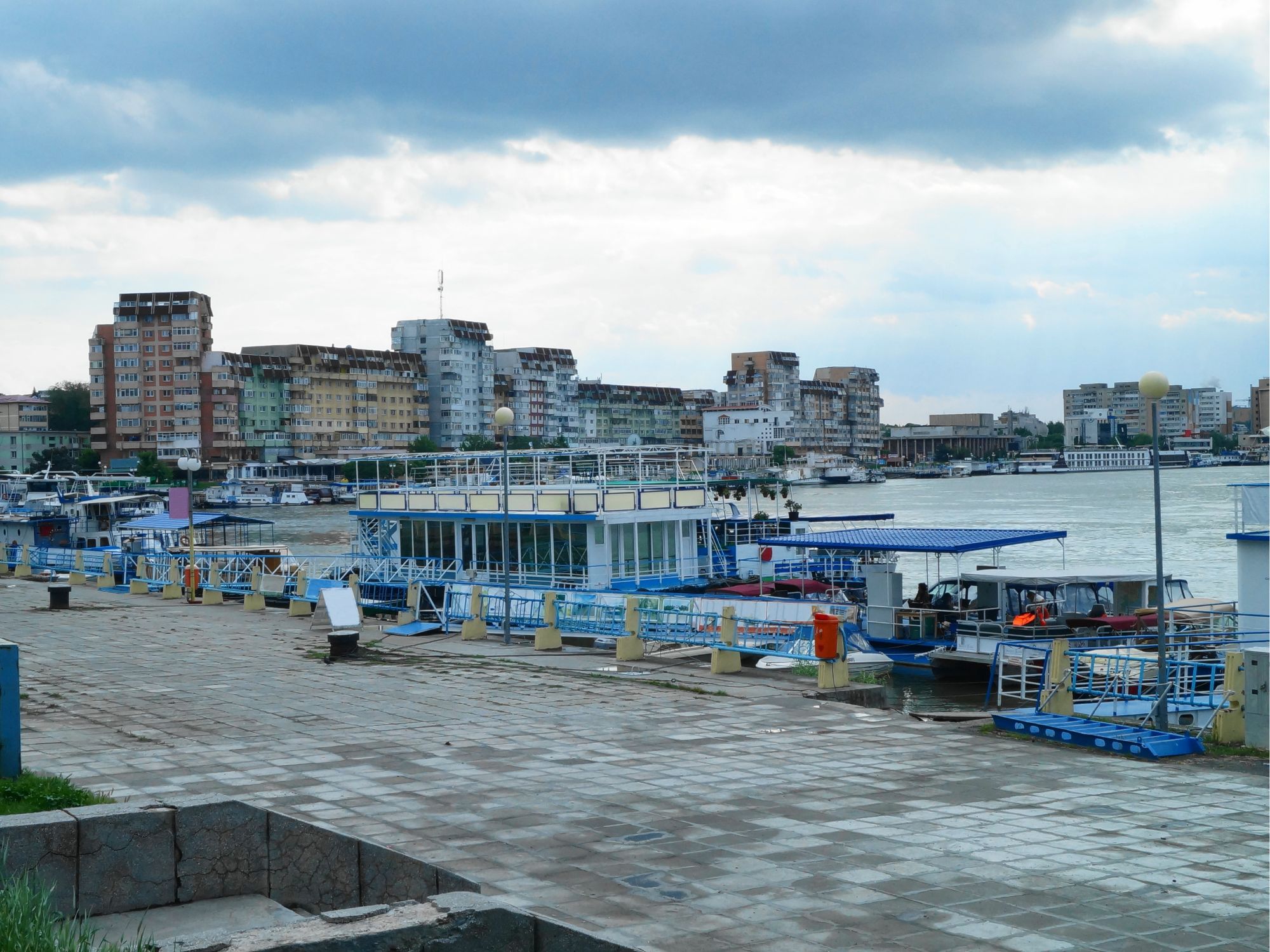 Boats docked at a bustling port in Tulcea, Romania, with modern buildings under a cloudy sky in the background.