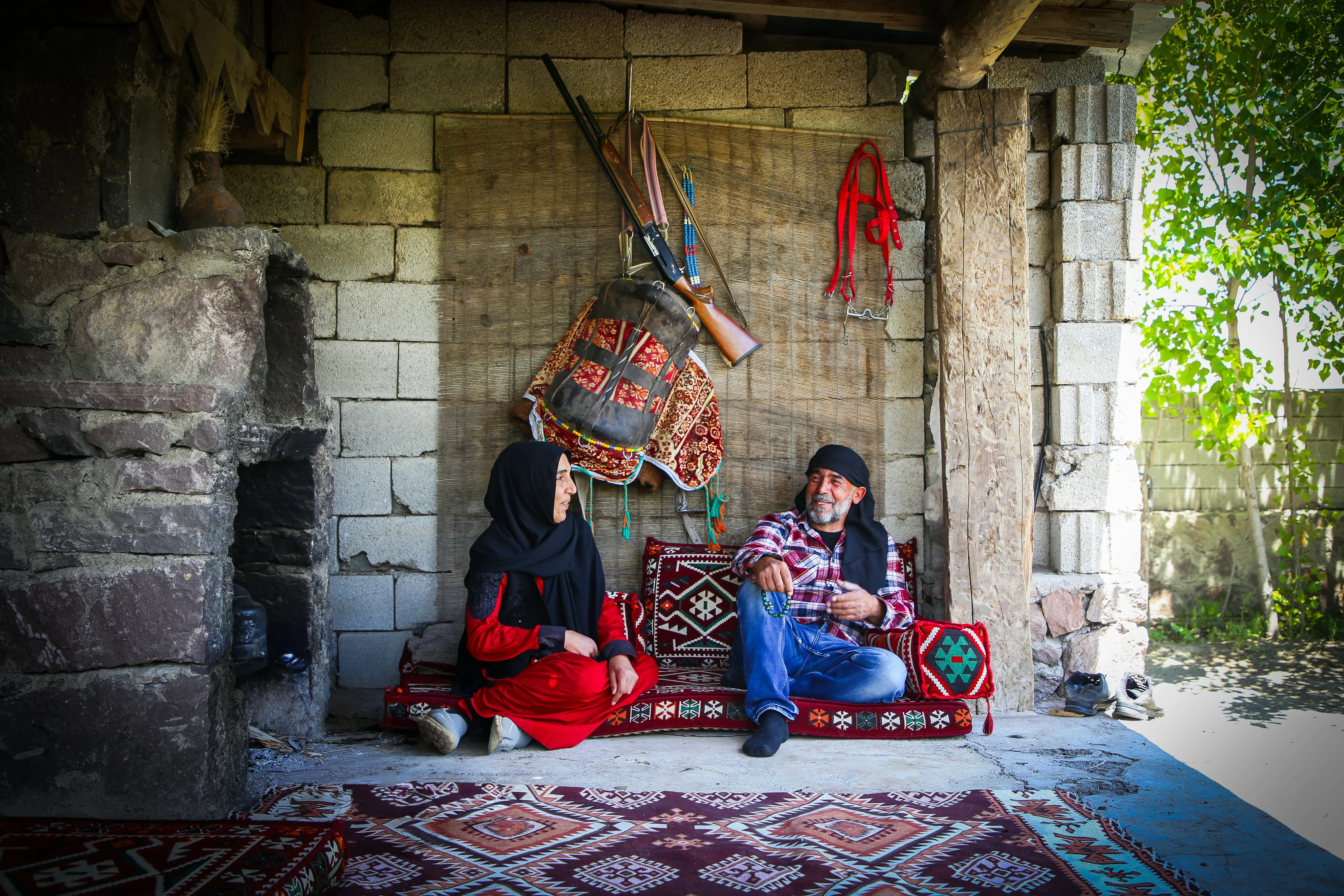 A Kurdish couple sitting on traditional carpets in a rustic home, engaged in conversation.