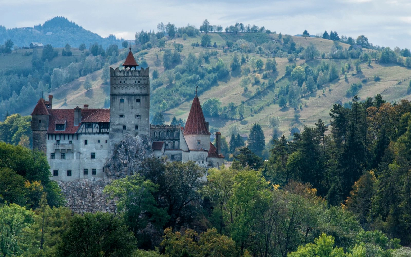 Bran Castle in Transylvania, with red roofs and stone towers, surrounded by green forests and rolling hills.