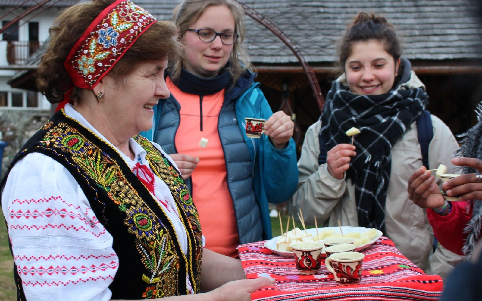 Local woman in traditional embroidered vest serves cheese and drinks to smiling hikers at a rural homestay in Romania.