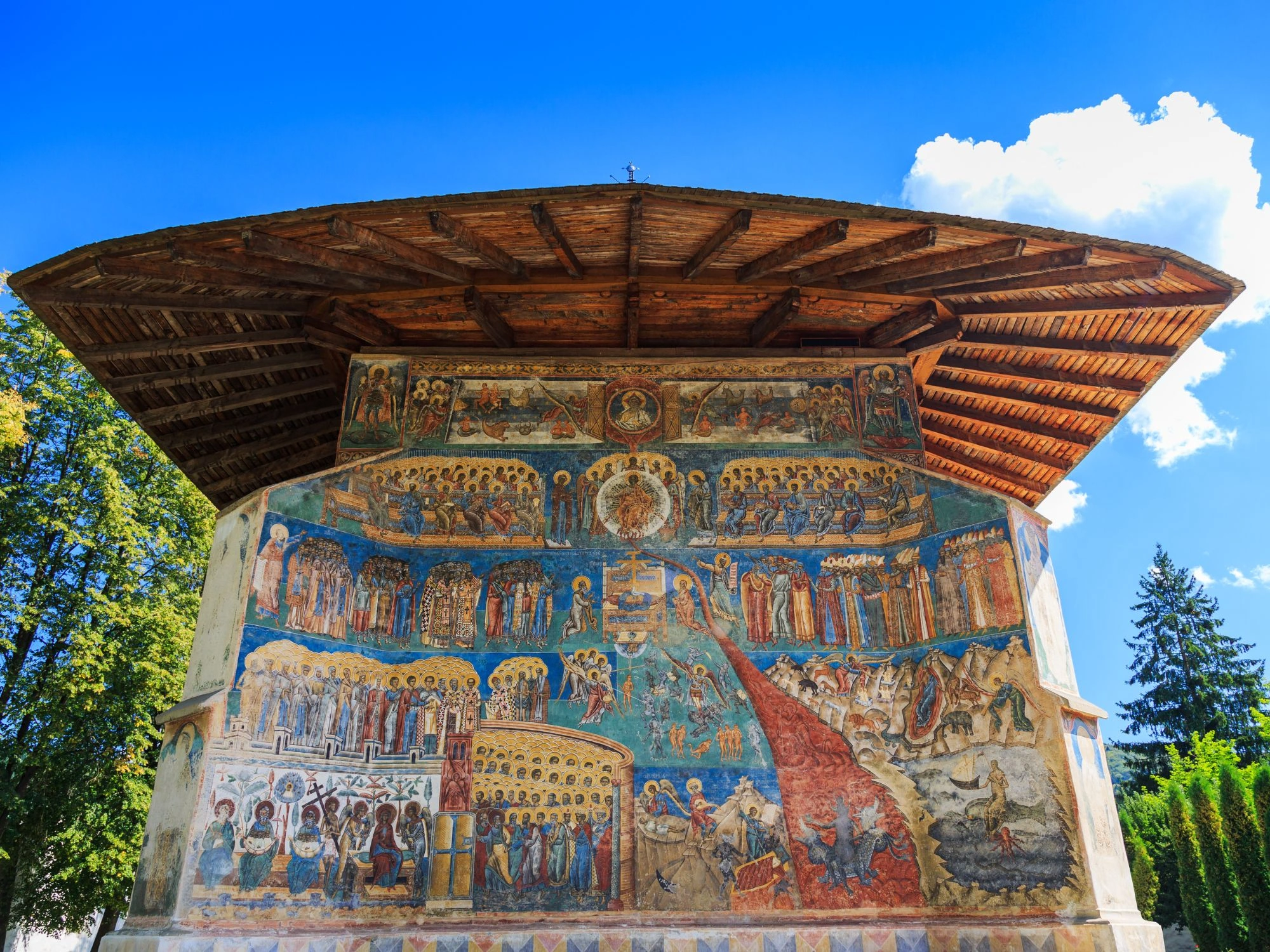 Painted monastery exterior with colorful biblical frescoes under a wooden roof.