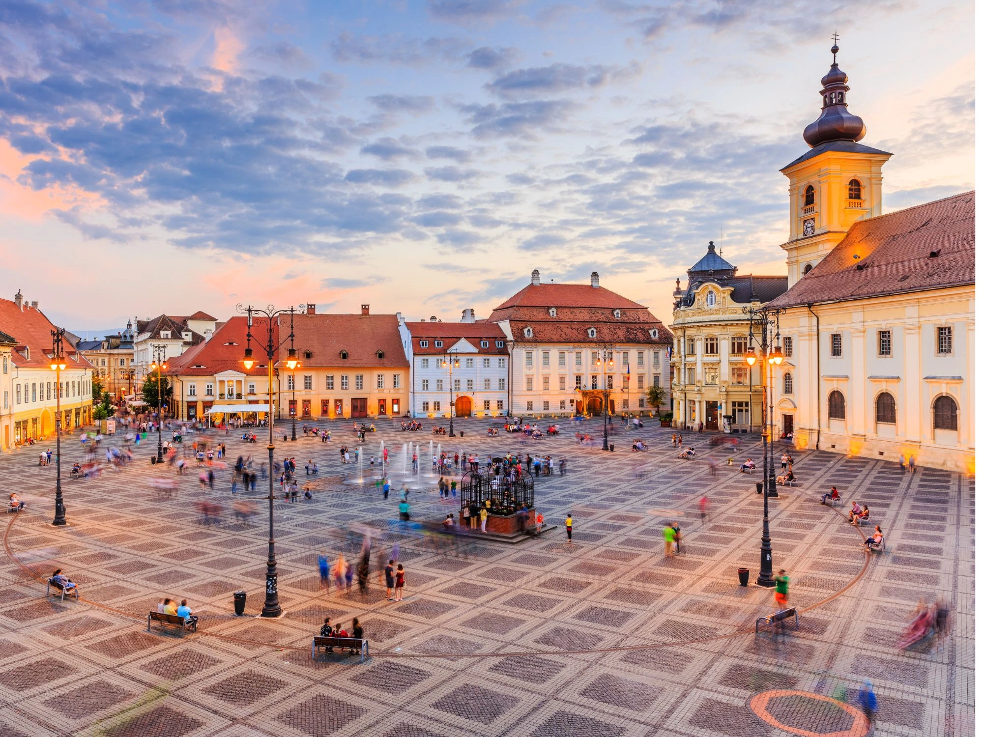People walking through Grand Square in Sibiu, Romania at sunset with pastel baroque buildings