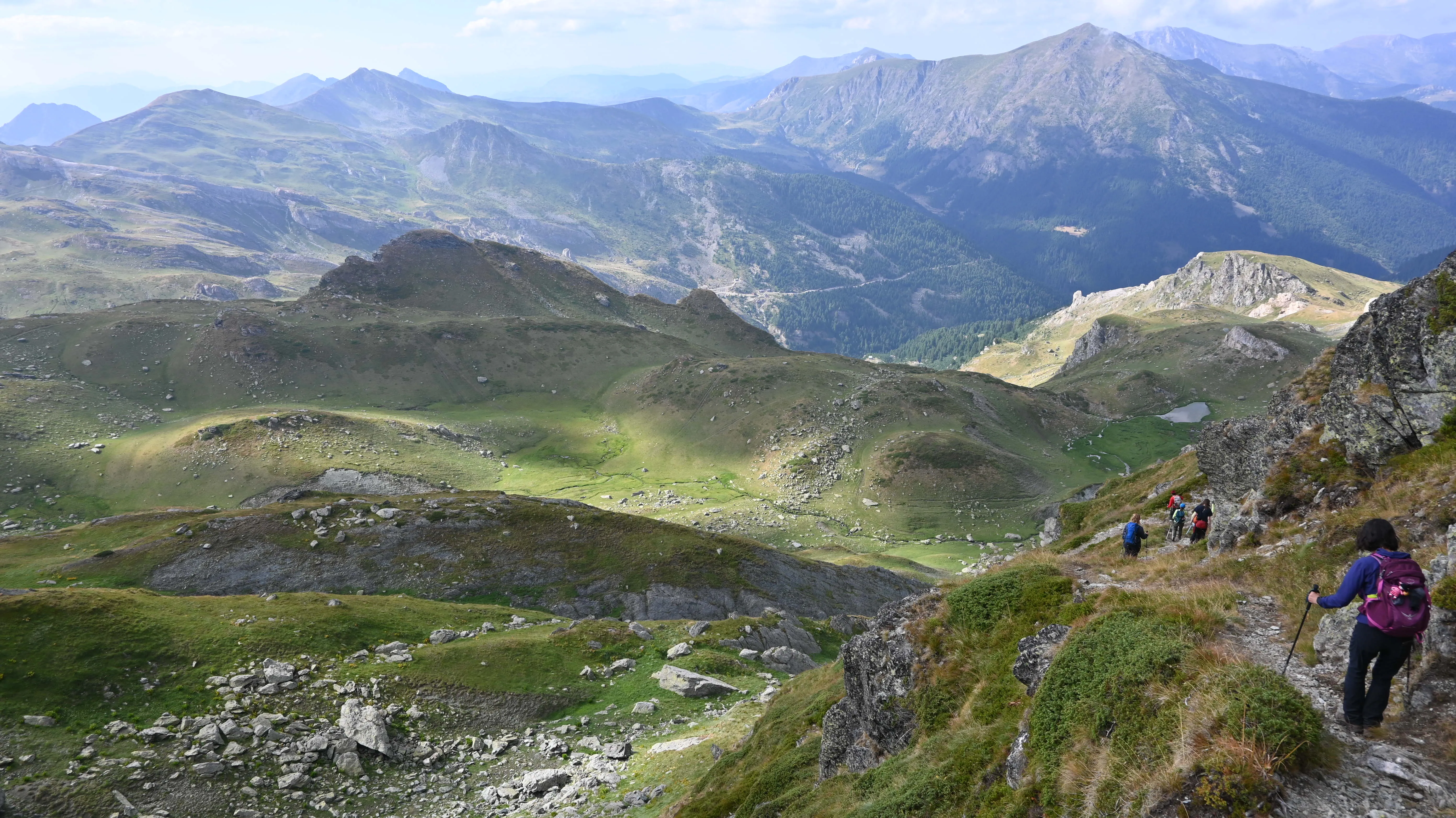 Hikers trekking down a green ridge on the Via Dinarica trail in Kosovo, surrounded by mountain views.