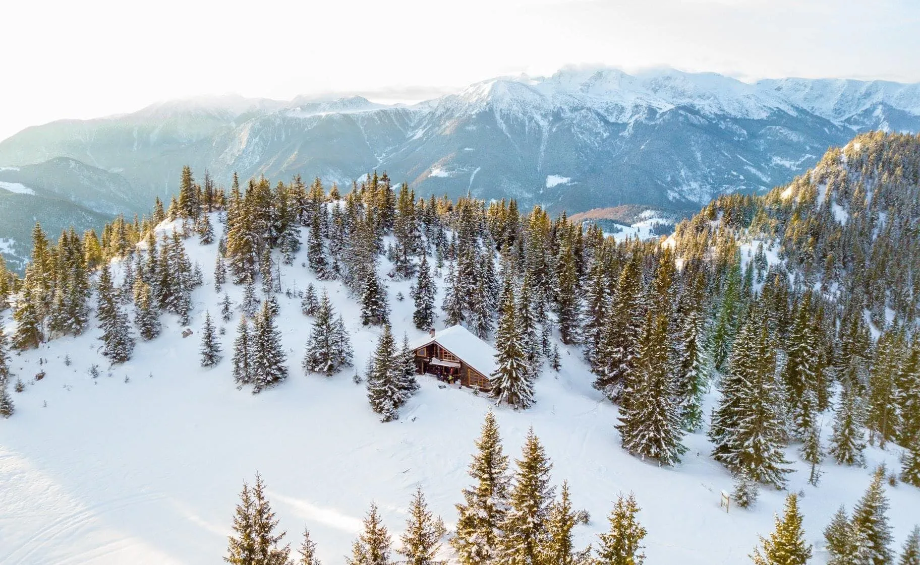 A snow-covered cabin nestled among evergreen trees with scenic views of the snow-capped Hajla mountains